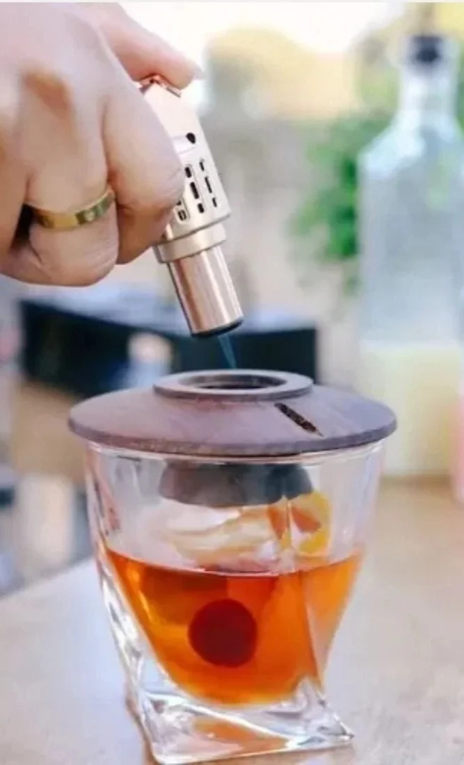 Hand holding a smoking gun-shaped device pointed at a glass container with liquid and floating objects on a table indoors.