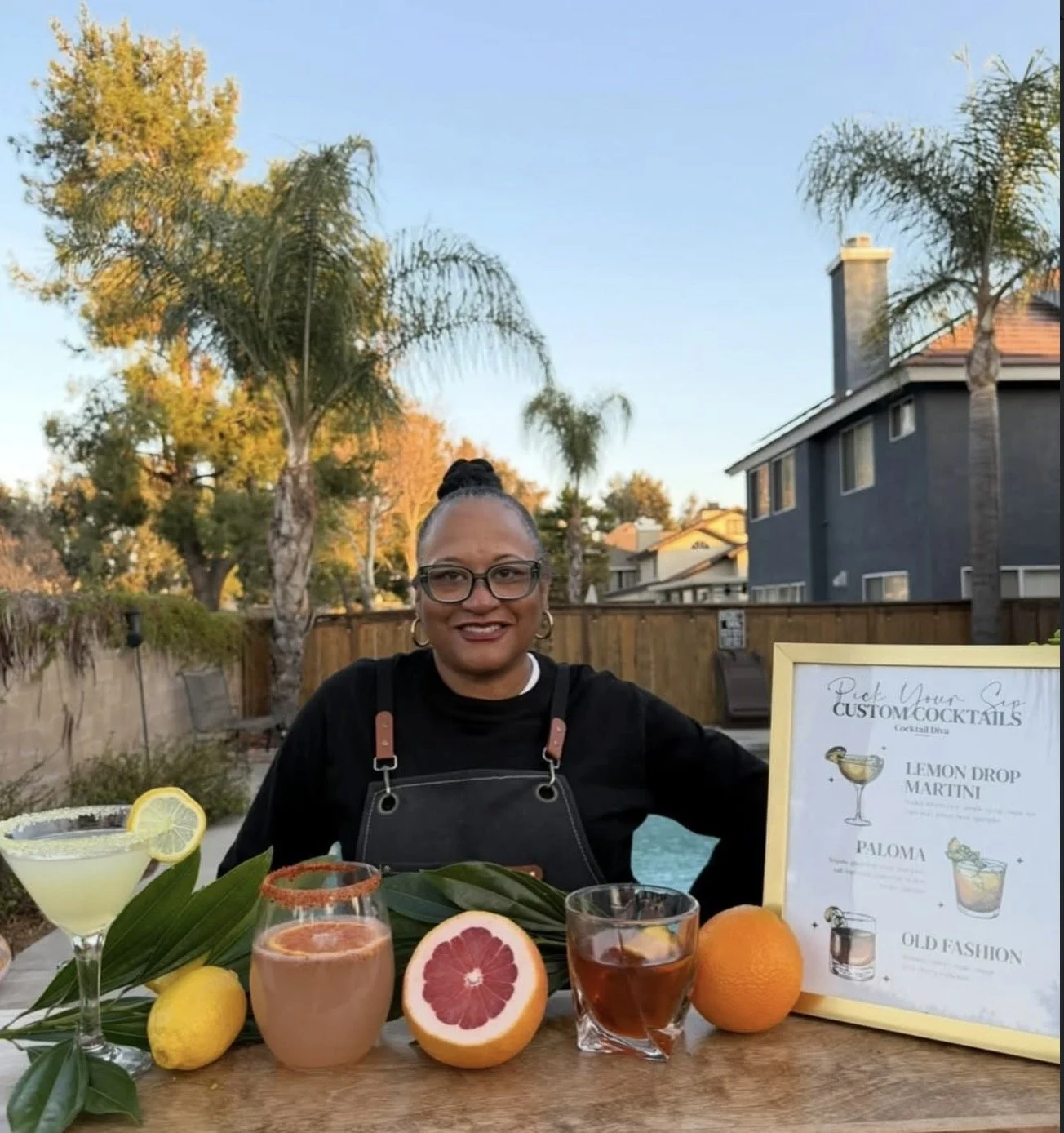 Woman smiling at a table with colorful cocktails, citrus fruits, and a menu for custom cocktails outdoors in a backyard with palm trees and houses.