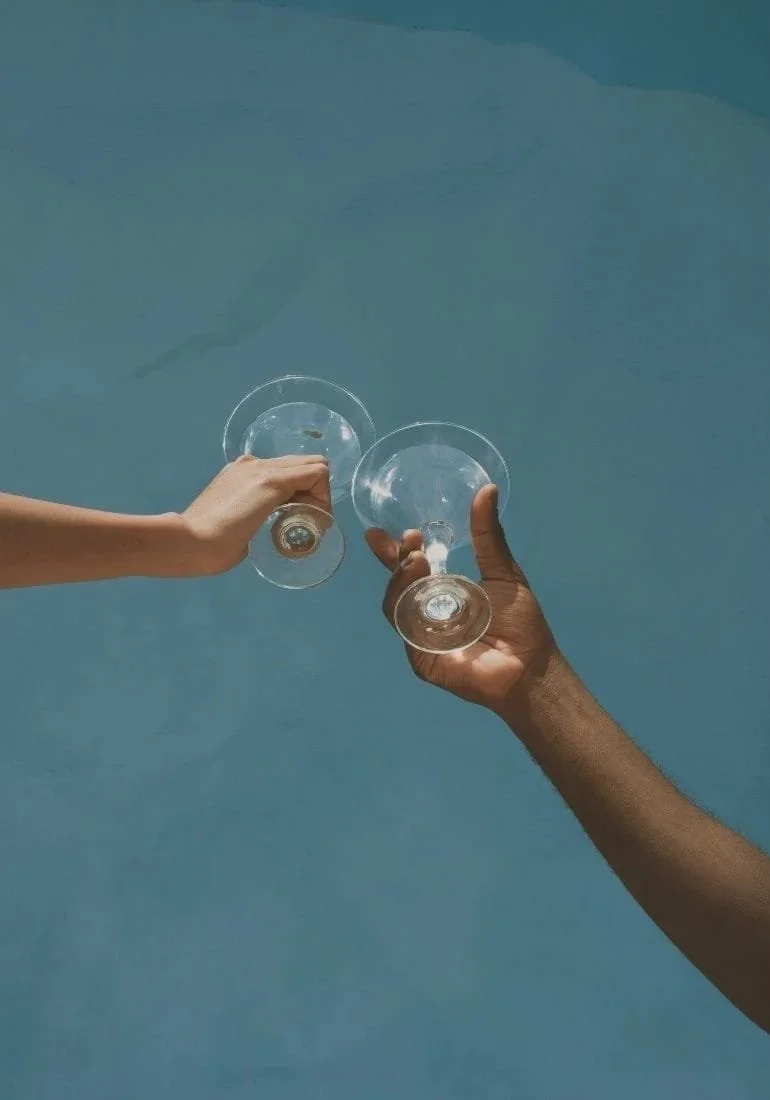 Two people are holding wine glasses, clinking them together over a pool of blue water.