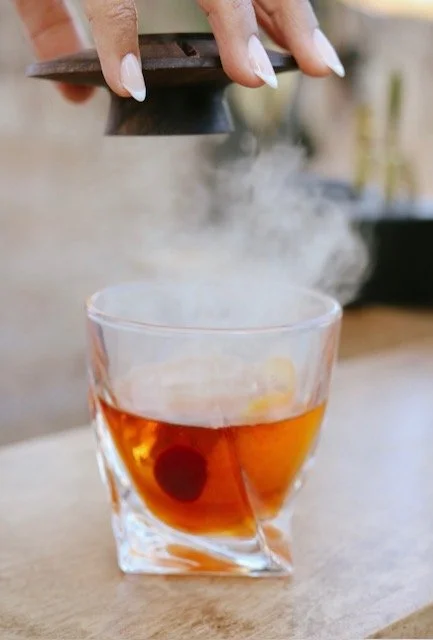 A hand holding a metal object above a glass of tea, with steam or vapor rising from the glass.