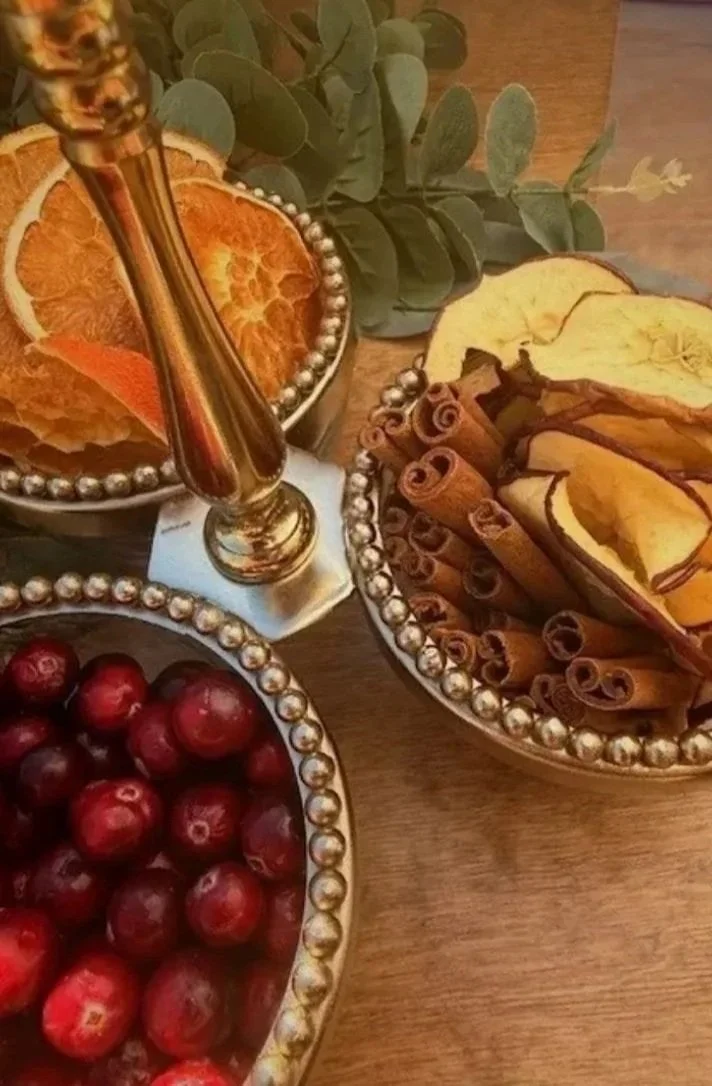 Decorative bowls of dried orange slices, cinnamon sticks, and red cranberries, with a gold candle holder and eucalyptus leaves on a wooden surface.