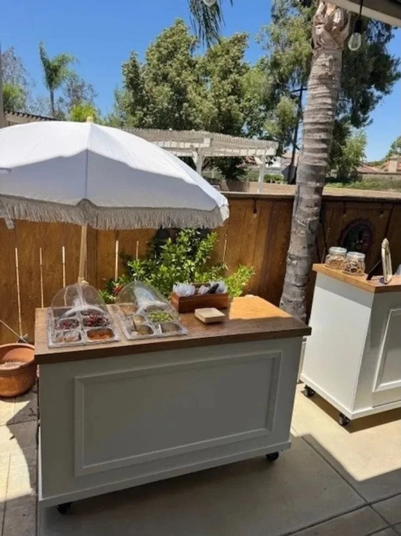 Outdoor food cart with a white umbrella, herbs, and small containers of food on a cart in a backyard with a wooden fence, trees, and a palm tree.