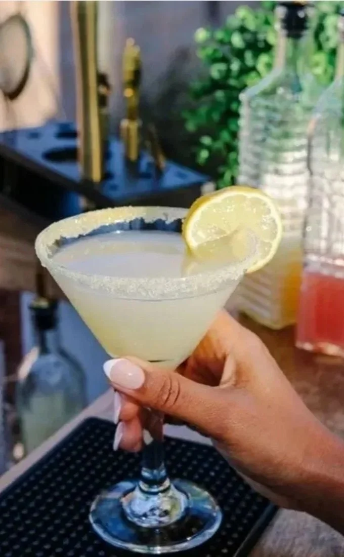 A person's hand holding a cocktail glass with a salted rim, a lemon slice garnish, and a pale yellow drink inside, on a bar counter.