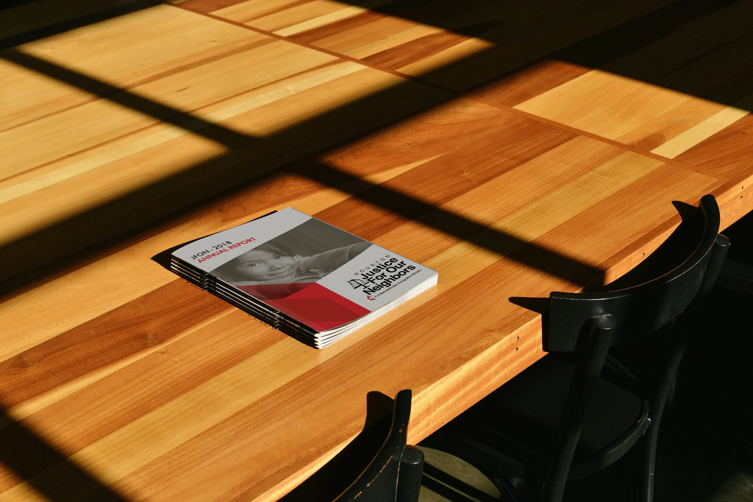 A wooden conference table with a stack of annual report booklets titled 'Justice for Our Neighbors' and 'Annual Report 2018'. There are two black chairs around the table, and the image is taken through a metal grate or rack.