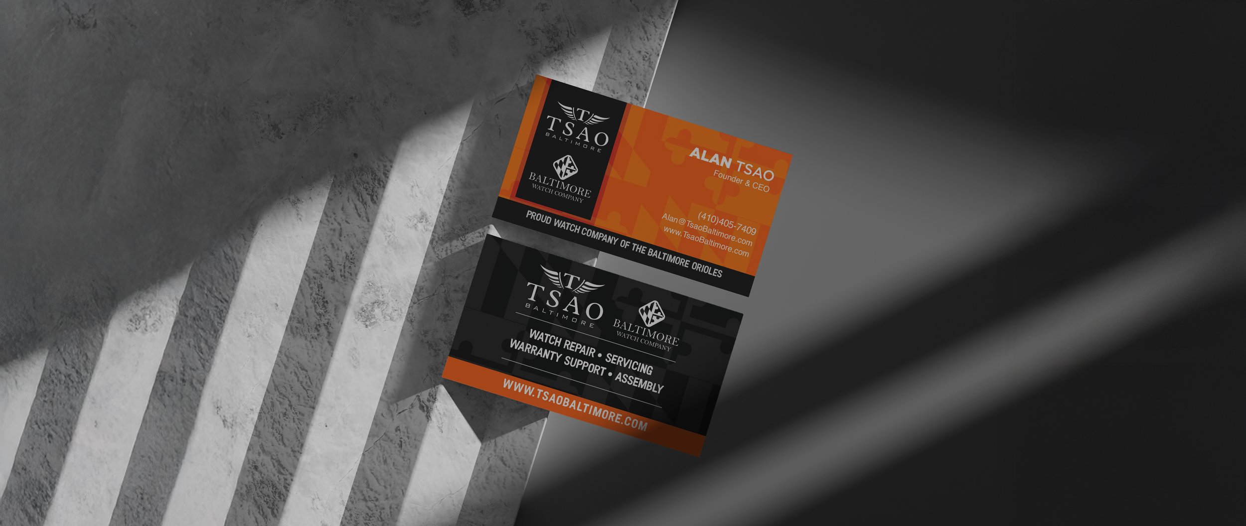 Two business cards for TSAO Baltimore, one orange and black, the other black and orange, placed on a textured gray surface, with shadows cast across the scene.
