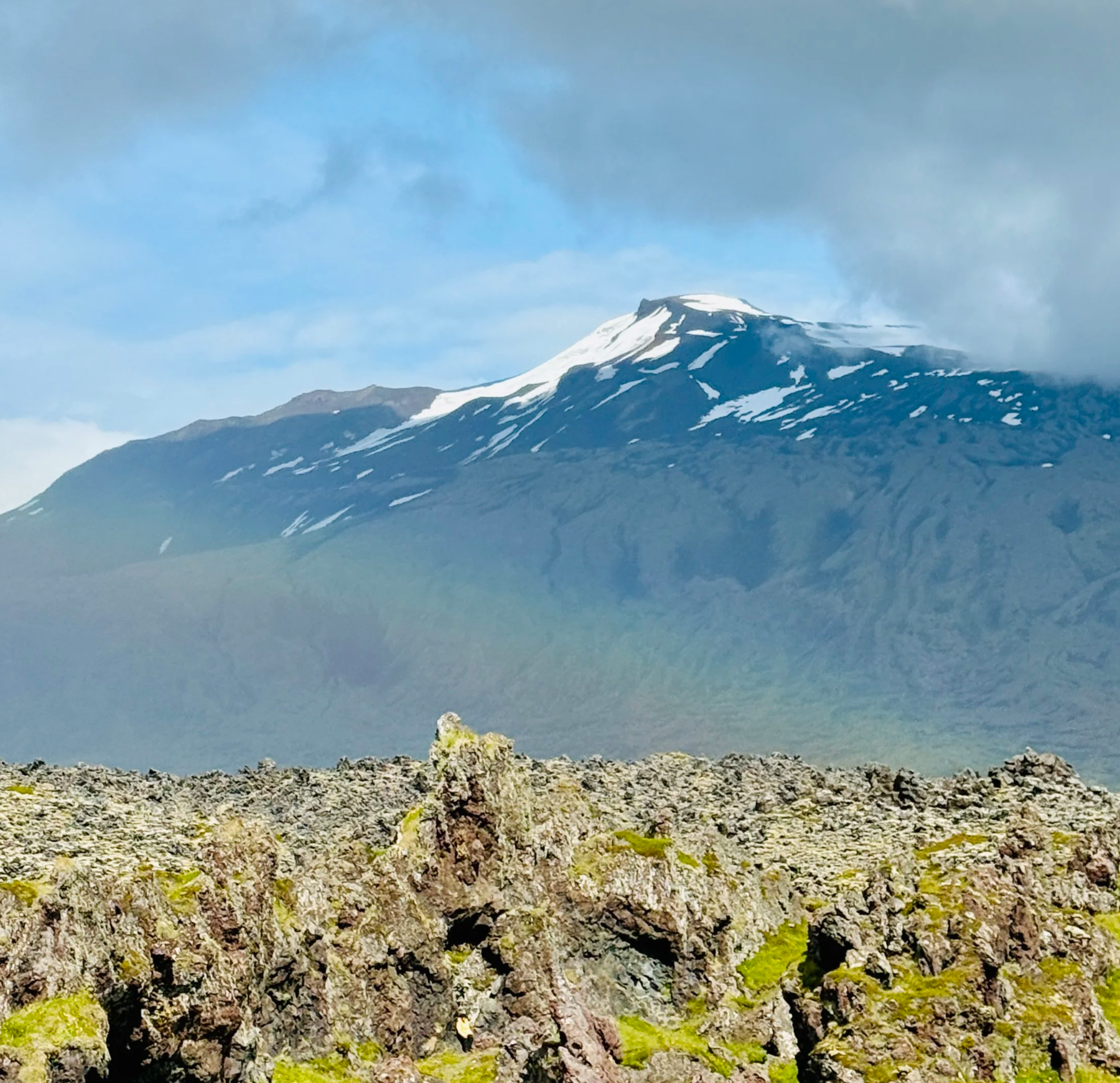 A snow-capped mountain with rugged rocky foreground and cloudy sky.