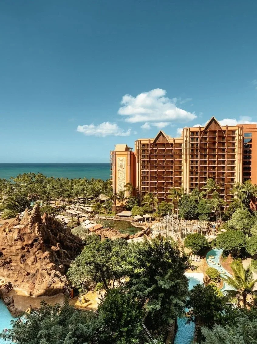 A large hotel with multiple floors and balconies situated near a tropical beach with palm trees and a water park with pools and water slides.