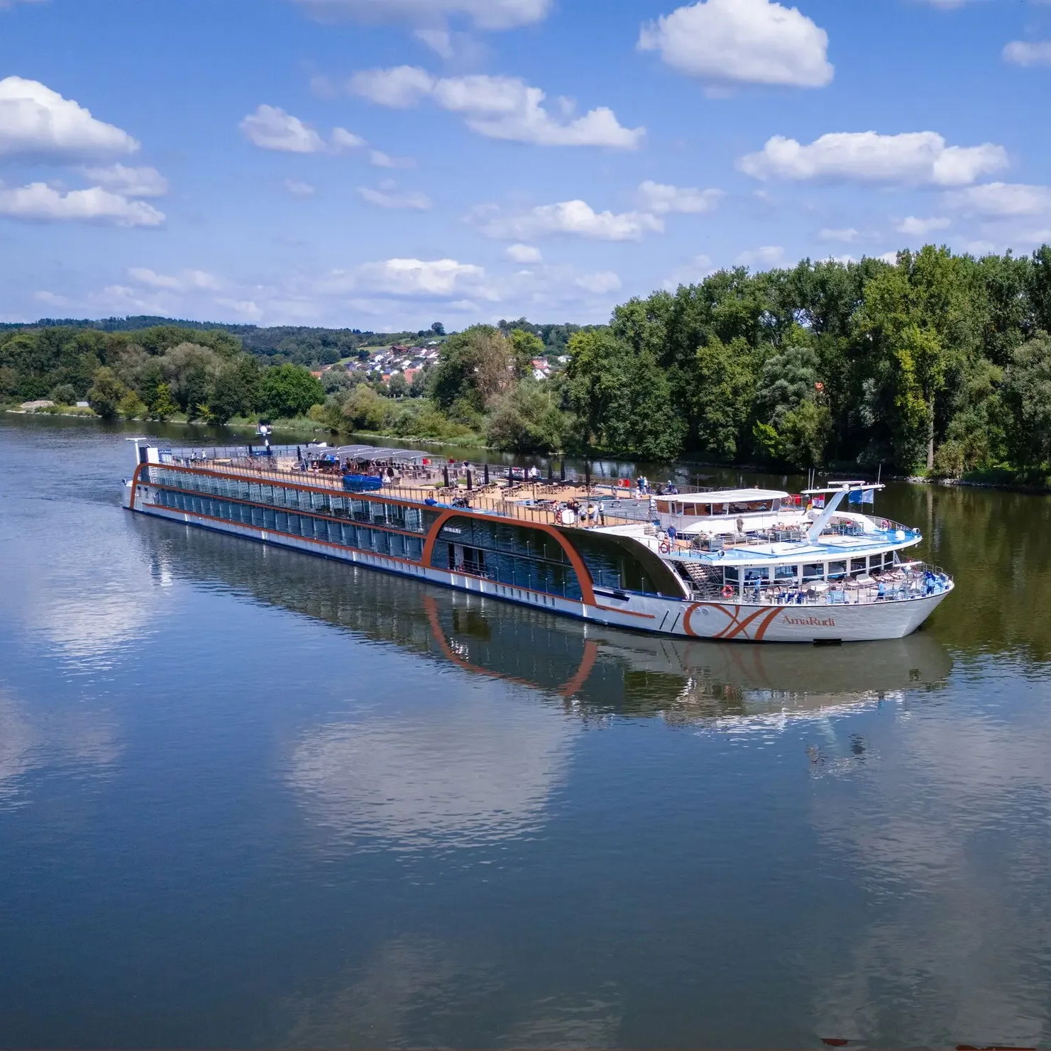 A large white and orange passenger cruise boat named 'Amal Rudi' is sailing on a calm river under a partly cloudy blue sky, with green trees and some houses visible on the riverbank in the background.
