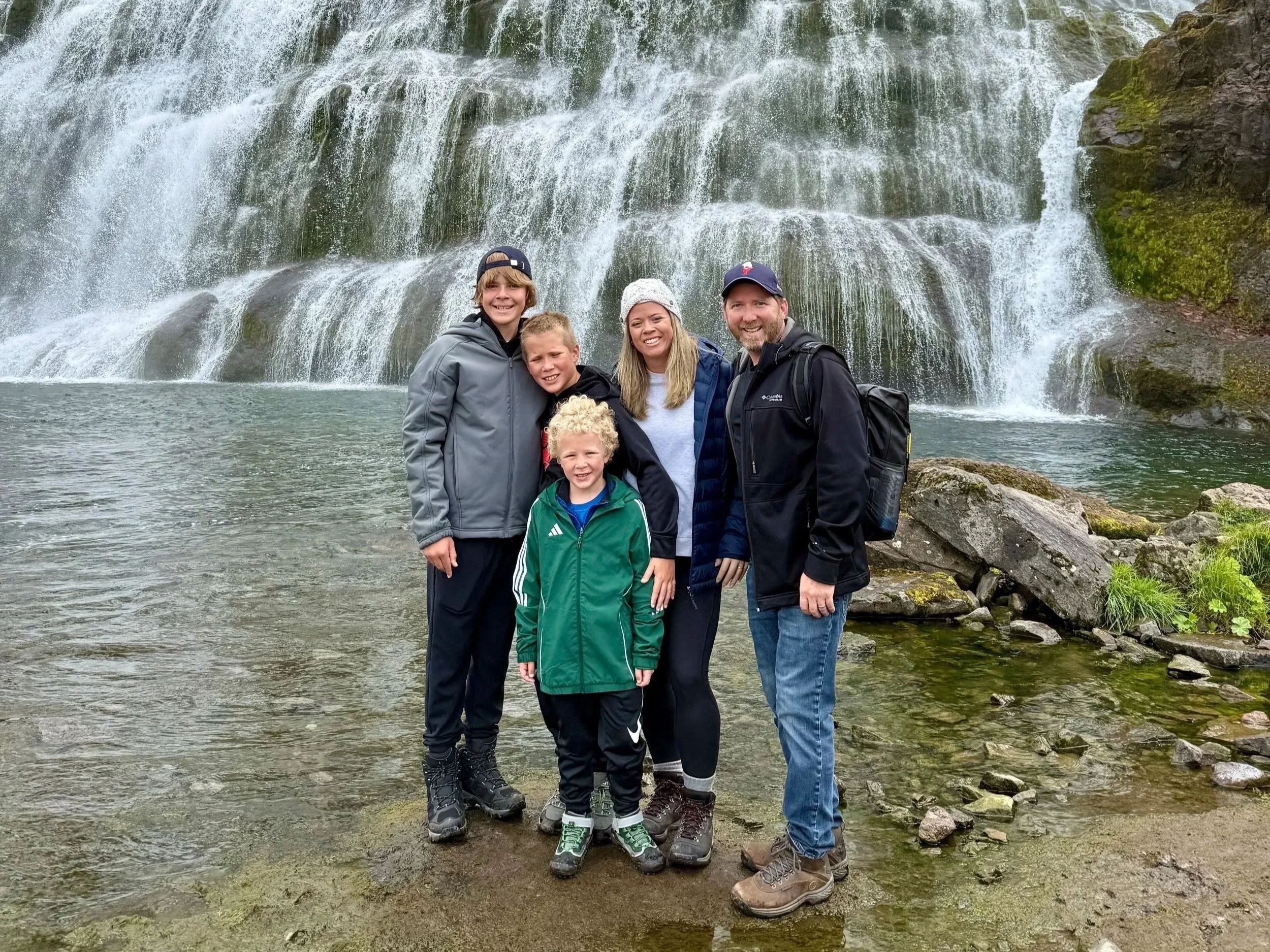 A family of five standing in front of a waterfall. The family includes two young boys, a young woman, a man, and a young girl. They are all smiling and dressed in outdoor clothing.