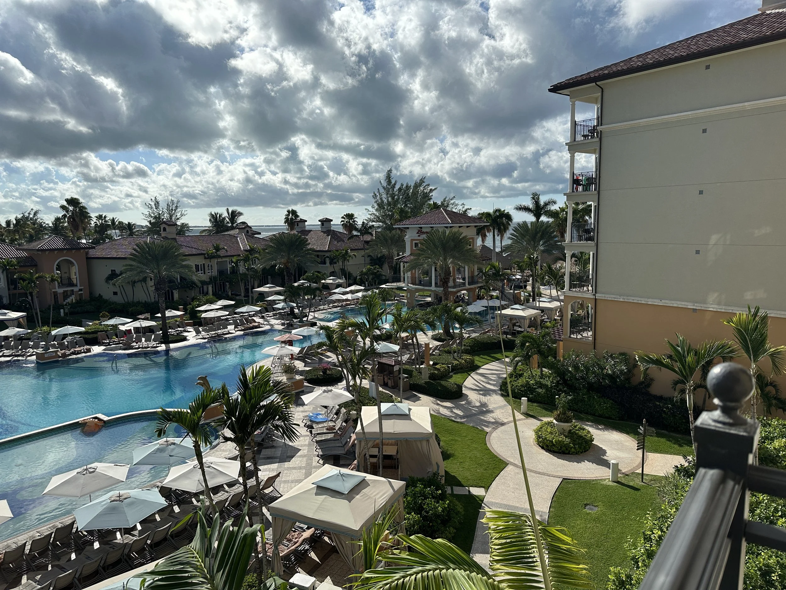 View of a resort pool area with lounge chairs and umbrellas, surrounded by palm trees and buildings under a cloudy sky.