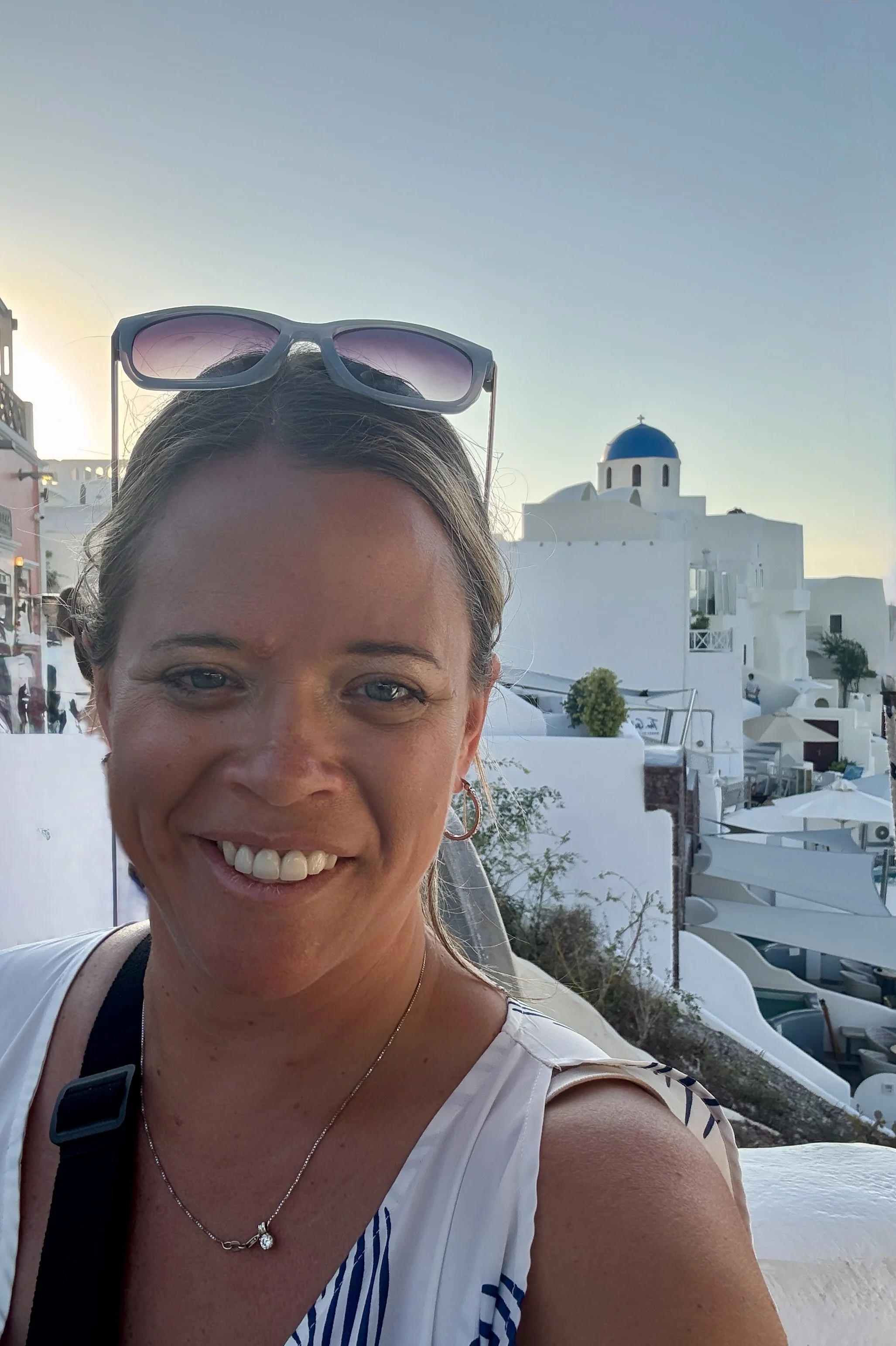 A woman smiling outdoors with white buildings and a blue-domed church in the background, likely in a Greek seaside town.