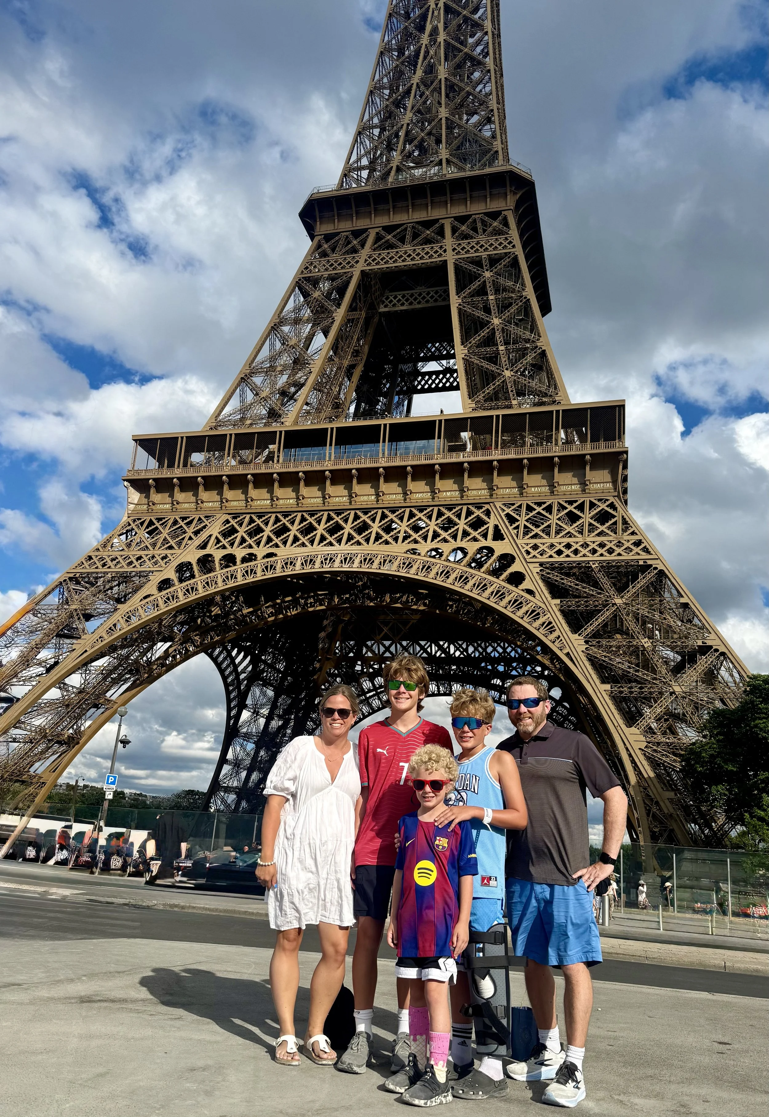 A family of six people standing in front of the Eiffel Tower in Paris, France. The group includes two adults and four children, all smiling and posing for the photo.