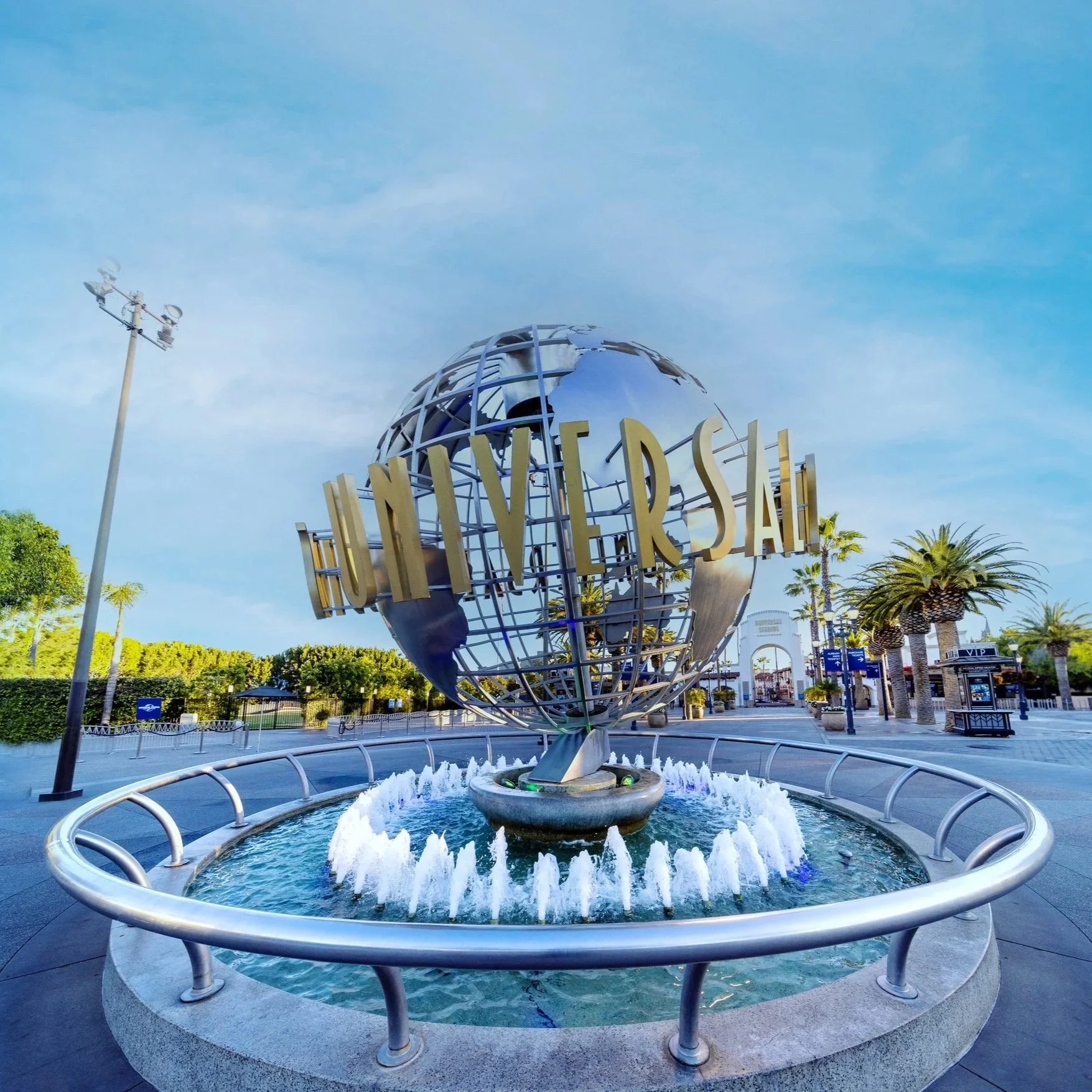 The Universal Studios globe statue in an outdoor area surrounded by a water fountain, with palm trees and a clear blue sky in the background.