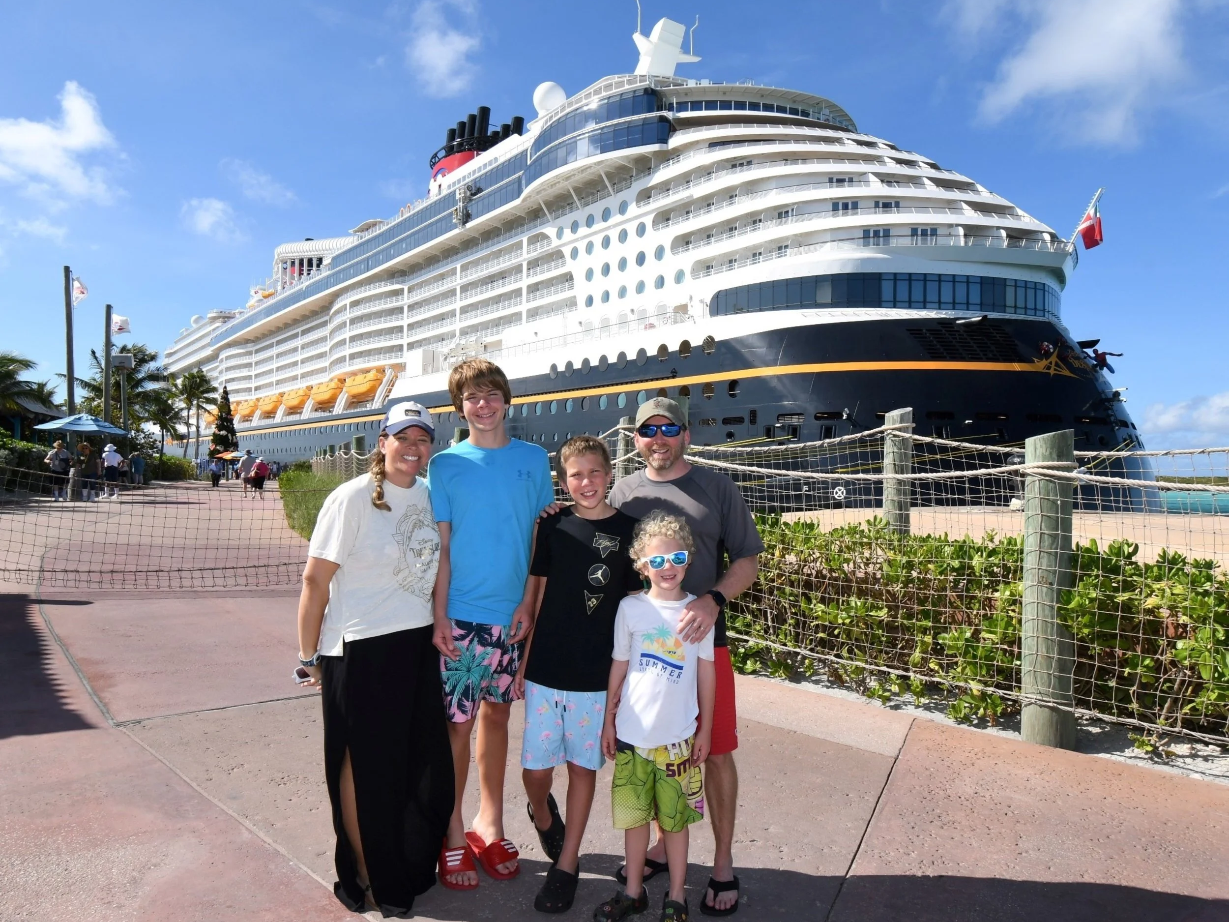 Six people standing in front of a large cruise ship, with a blue sky and some palm trees in the background.
