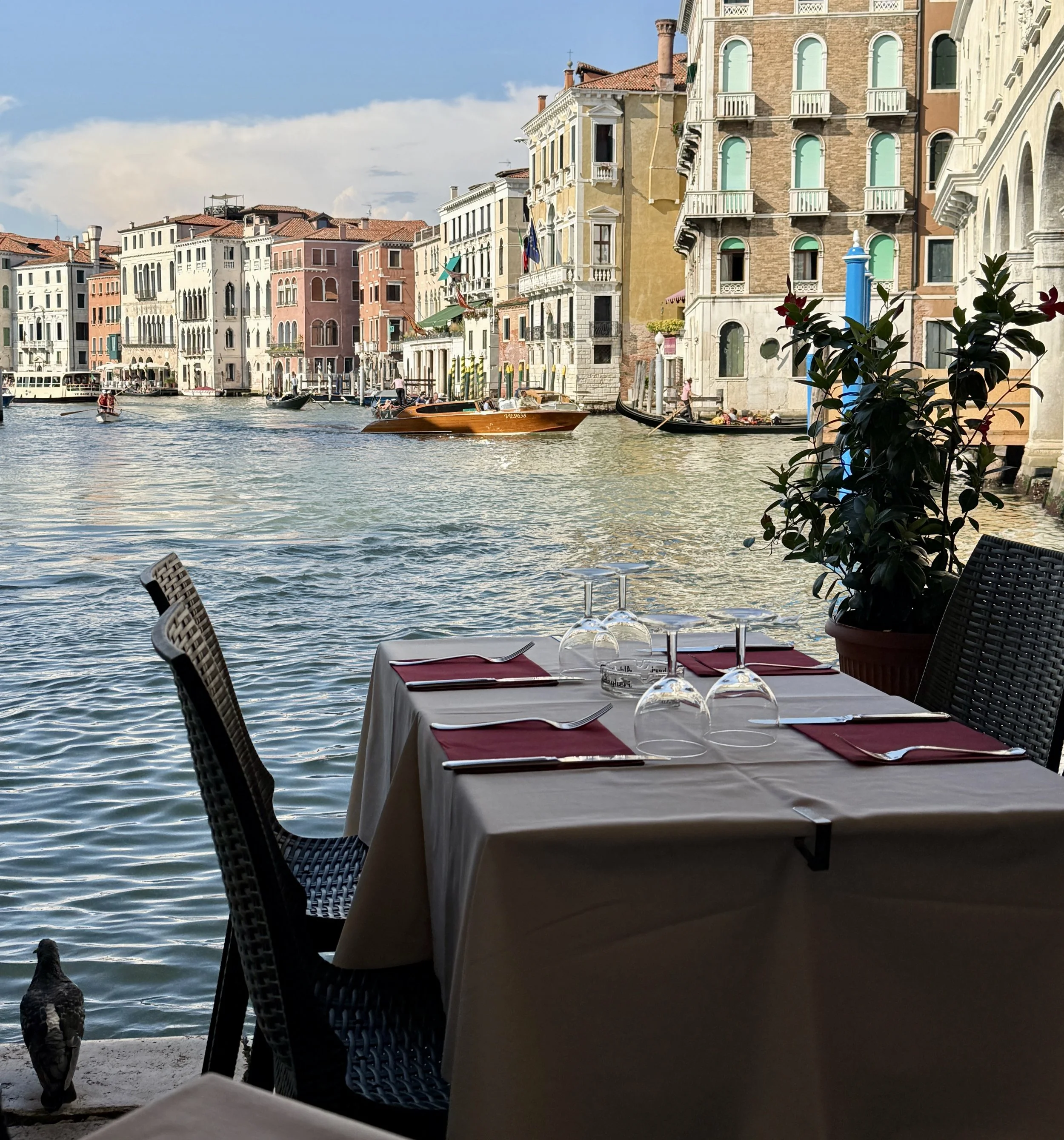 View of colorful buildings along a canal with boats docked, seen from a restaurant table with wine glasses and chairs in the foreground.