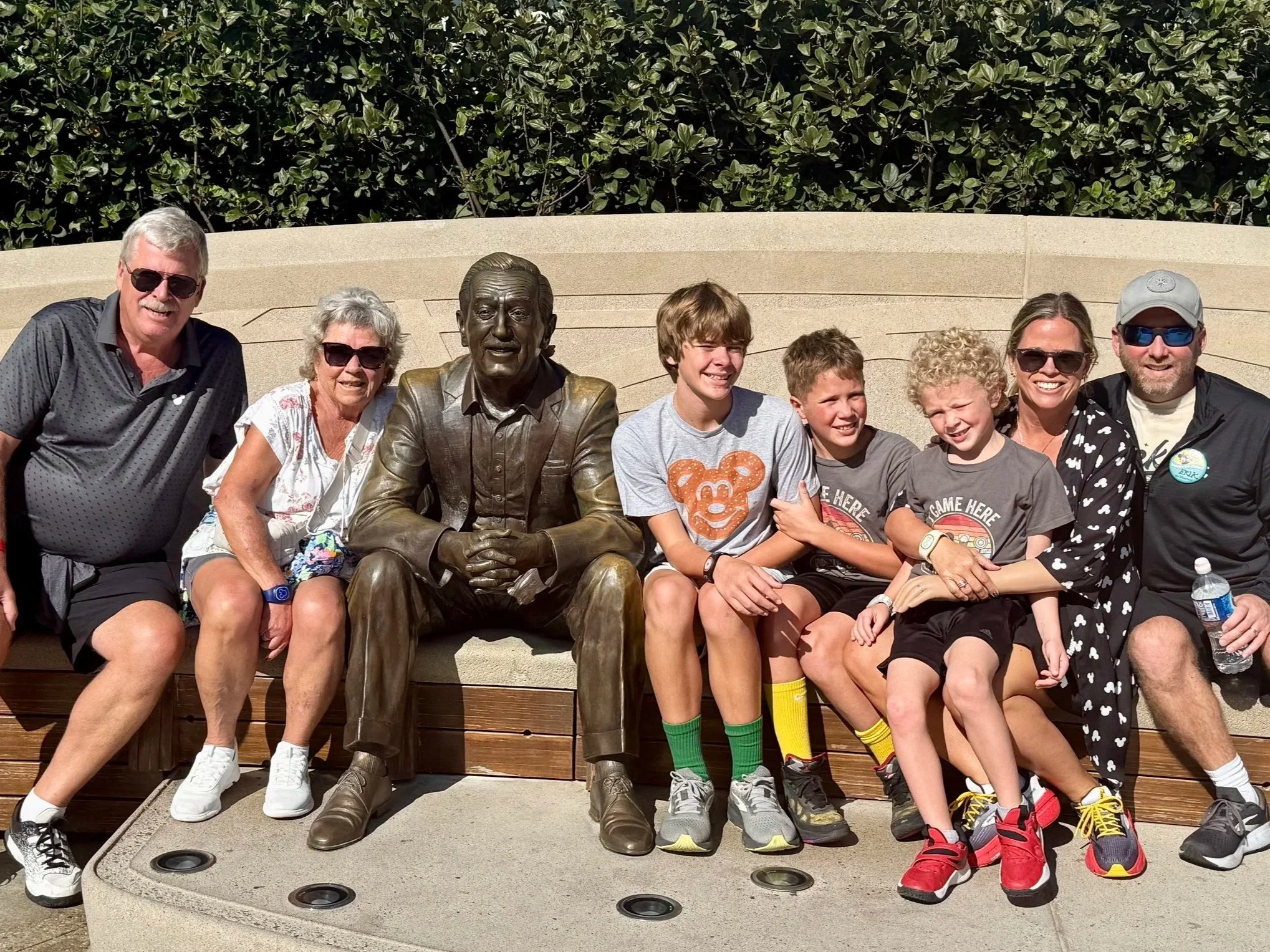 A group of people sitting on a bench with a statue of a man in the center. The group includes adults and children, all smiling. The background features green bushes and a beige wall. The people are wearing casual summer clothing and sunglasses, and some have water bottles.