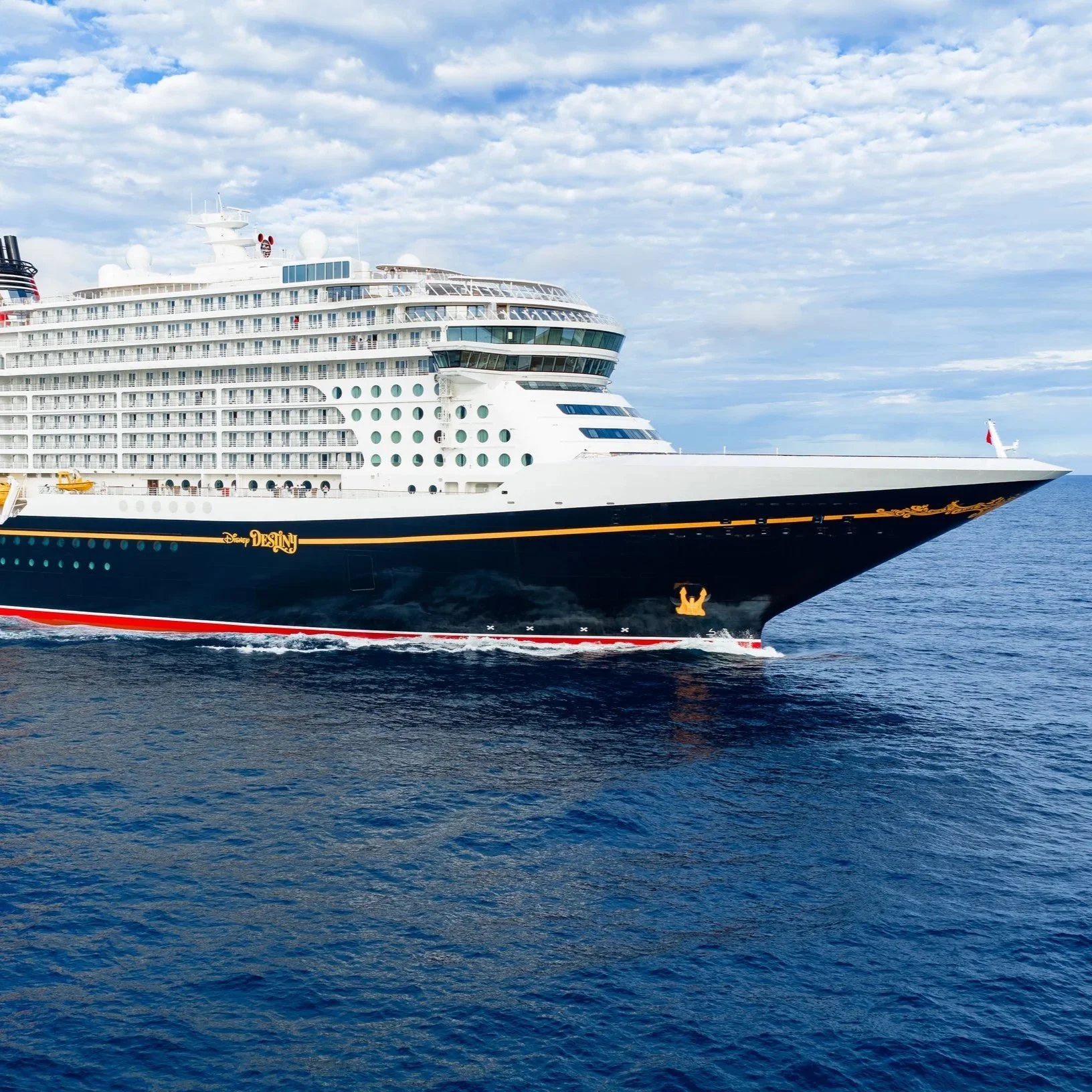 A large white cruise ship with multiple decks and balconies sailing on a calm open sea under a partly cloudy sky.