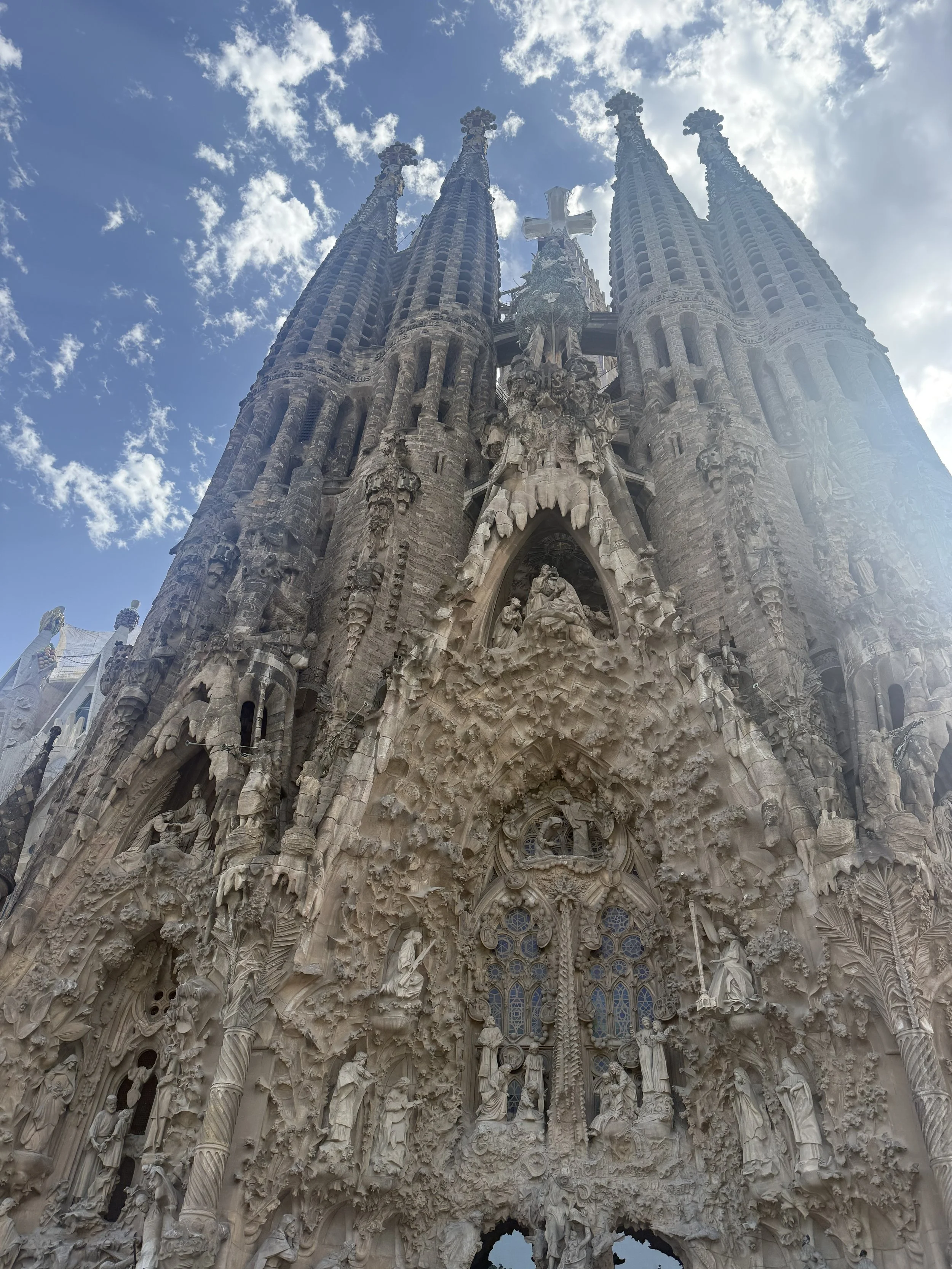 Looking up at the detailed facade of the Sagrada Familia, a large Catholic basilica in Barcelona, Spain, with ornate sculptures and tall towers under a partly cloudy sky.