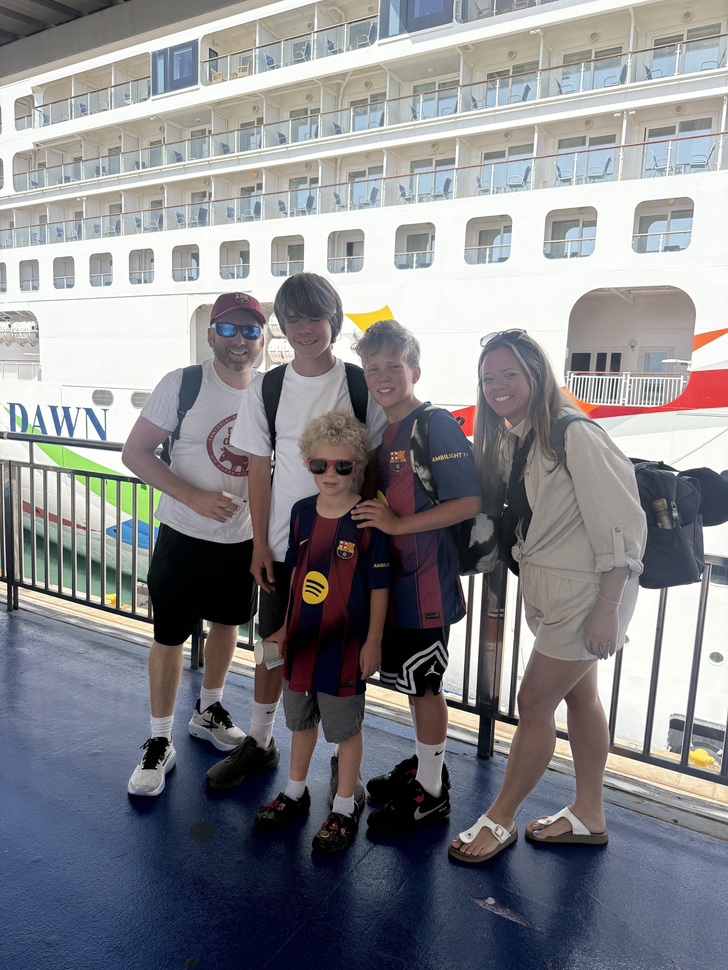Family standing on a ship's deck with a large cruise ship in the background, smiling for a photo.