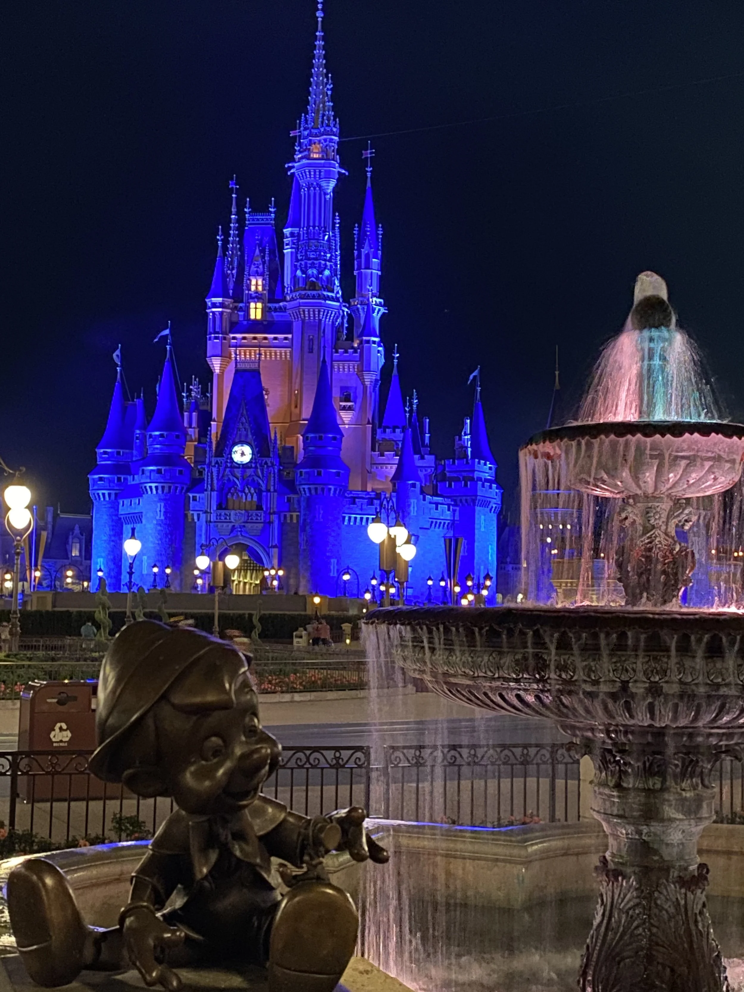 Night view of Cinderella's Castle illuminated in blue lights at a Disney theme park, with a decorative fountain and a statue of a young boy in the foreground.
