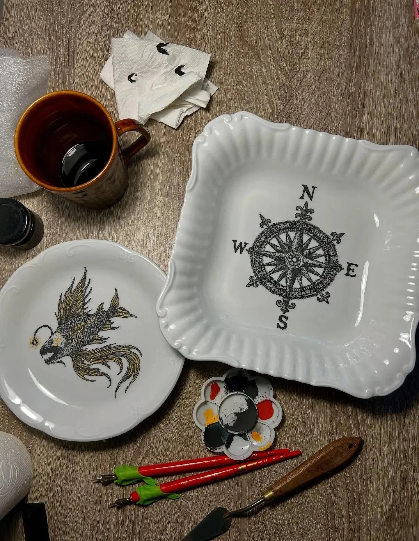 A table with art supplies, including a palette with black, red, and yellow paints, a ceramic square bowl with a compass design inside and a dragon illustration on a plate, a cup with a dark beverage, tissue paper, and various tools.