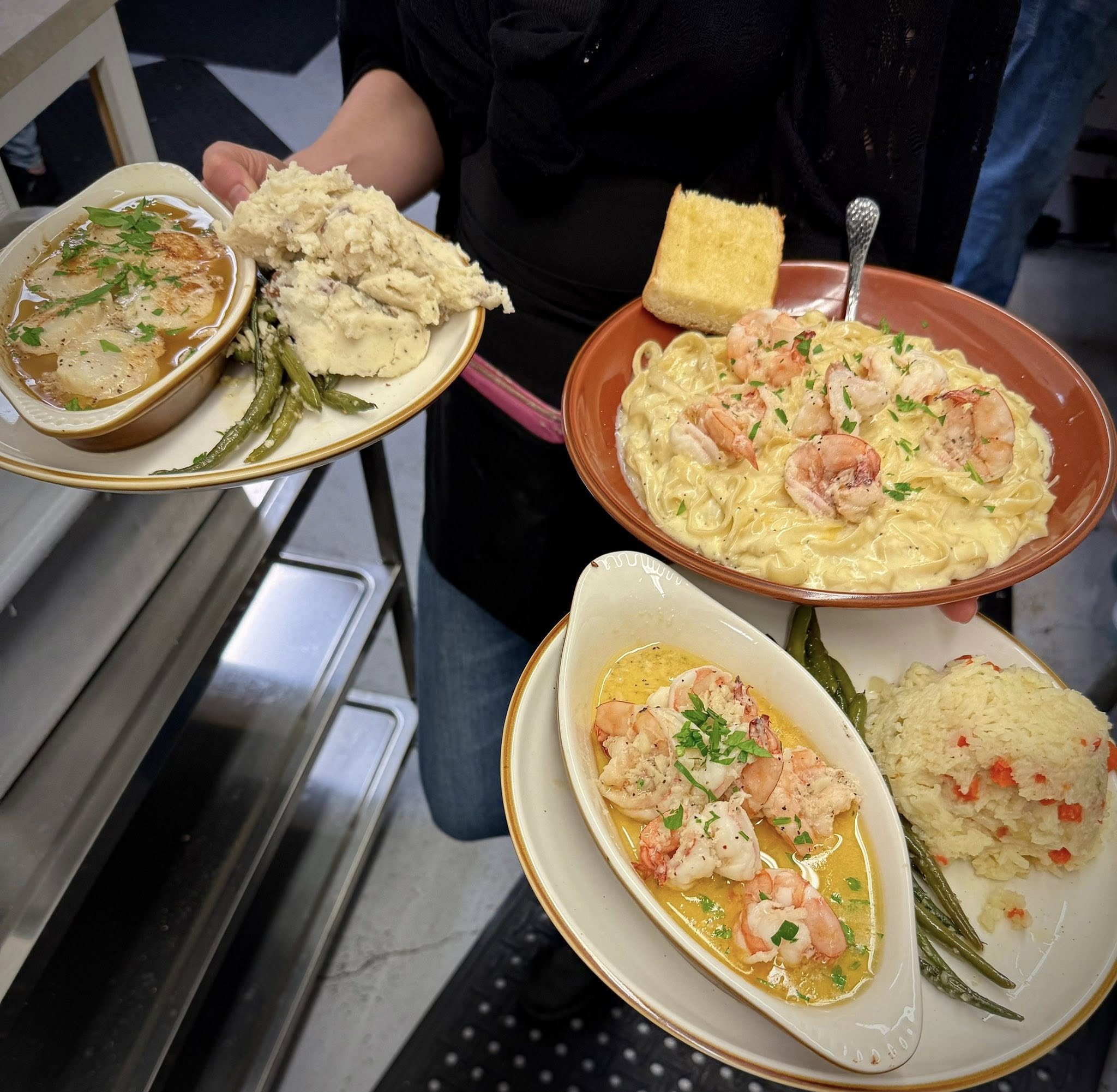 A person holding three plates of food, including shrimp pasta, mashed potatoes, green beans, and a bowl of soup.