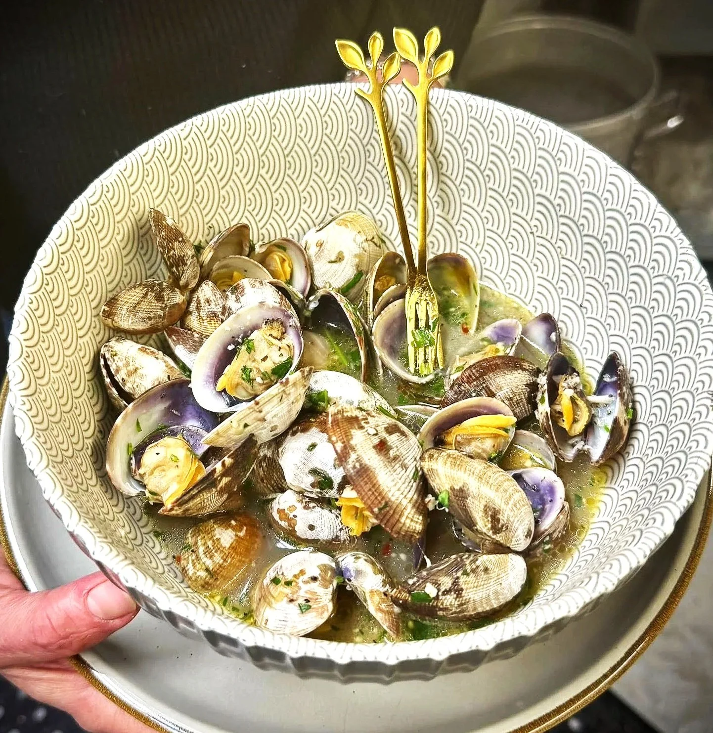 Bowl of cooked clams with green herbs and yellow seasoning, garnished with a gold fork and fork-shaped garnish, on a white patterned bowl.