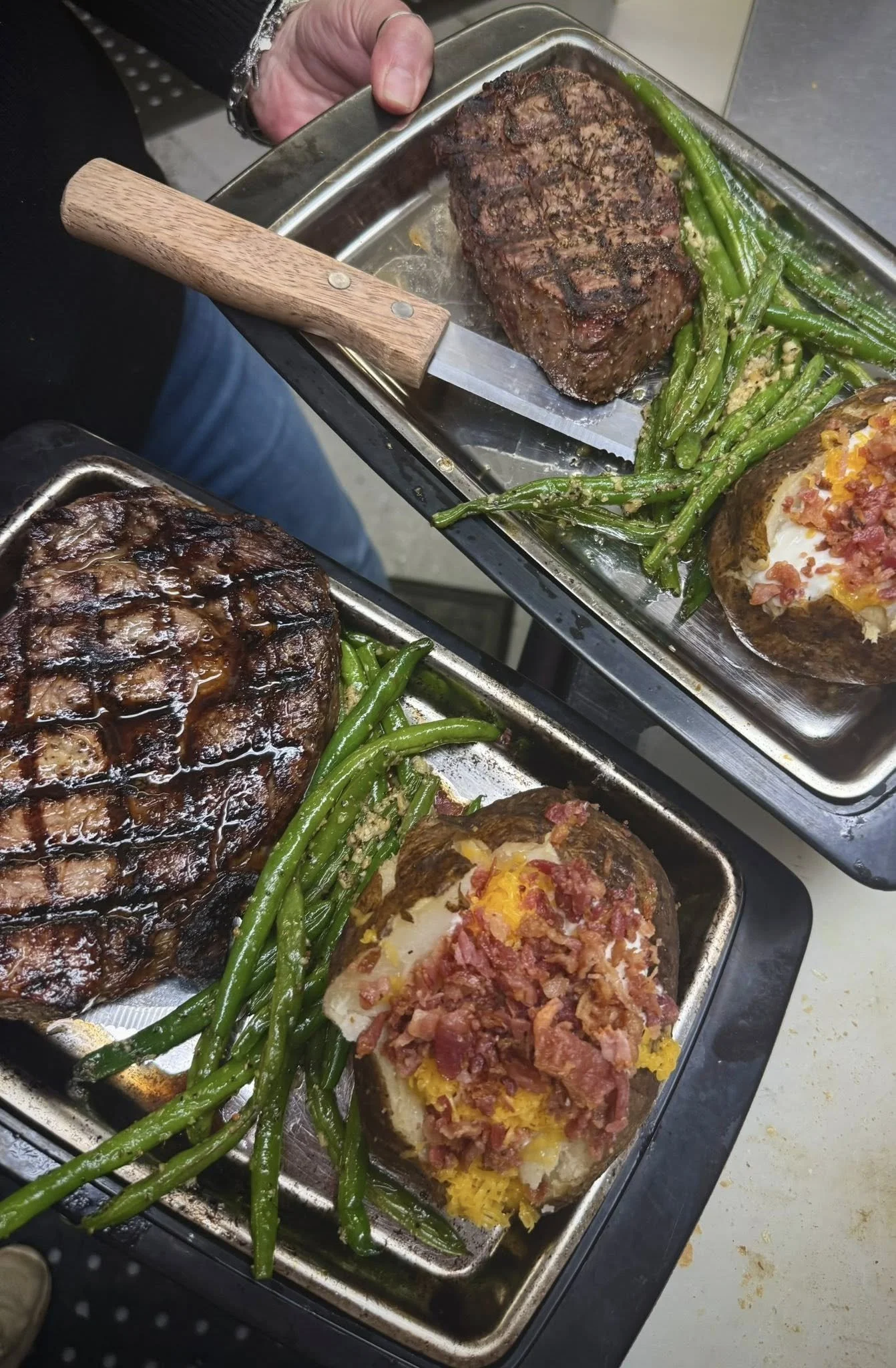 Two trays of cooked food including baked potatoes topped with bacon bits and shredded cheese, grilled steaks, and green beans.