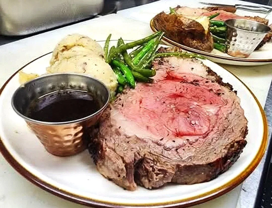 A plate of roasted prime rib with mashed potatoes, green beans, and a cup of au jus. In the background, there is another plate with cooked meat and a small metal cup.