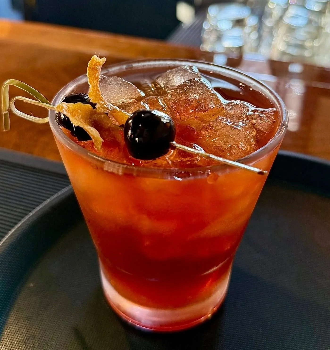 A manhatten cocktail with black cherries and lemon twist garnish on a black tray.