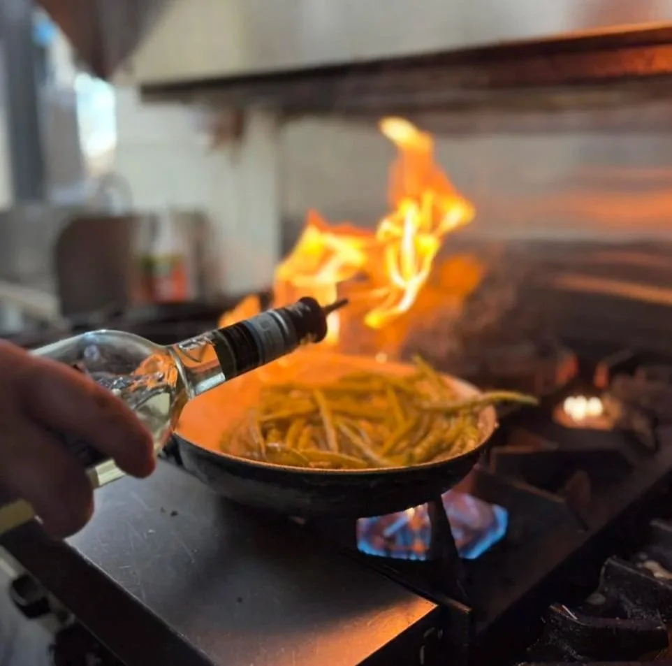 A person is pouring liquid from a bottle into a frying pan on a stove, causing a large flame to ignite.