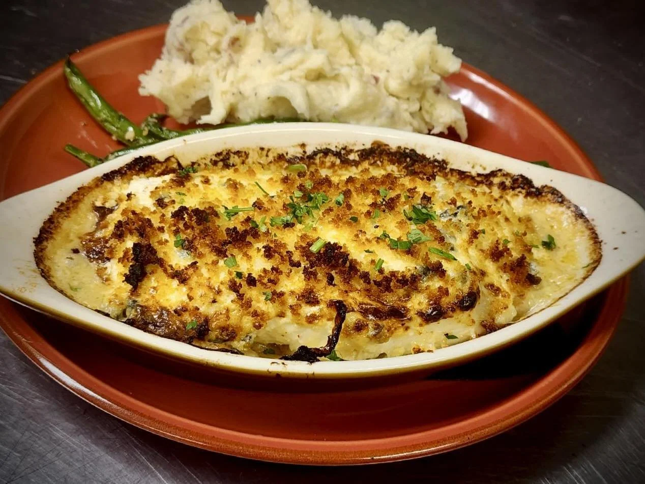 Baked casserole with herbs served in an oval dish, accompanied by mashed potatoes and green beans on a round brown plate.