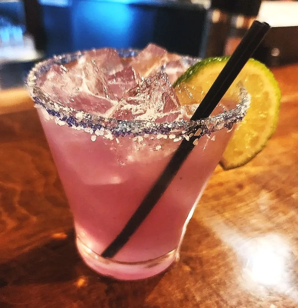 A Lavender Margarita cocktail with salt-rimmed glass, ice cubes, a slice of lime, and a lime wedge, with two black straws on a wooden bar counter.