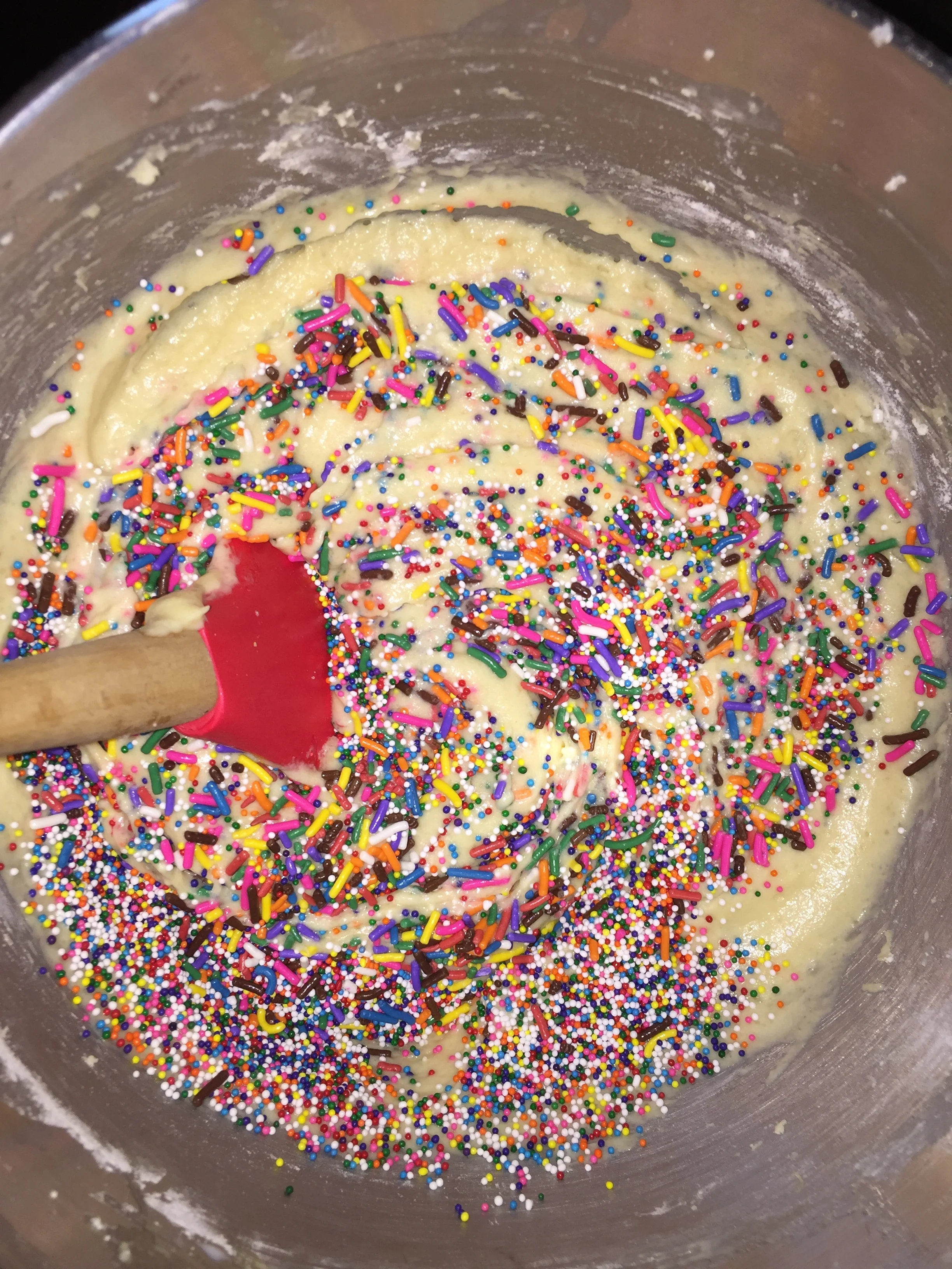 A mixing bowl containing yellow batter with colorful rainbow sprinkles and a red spatula.