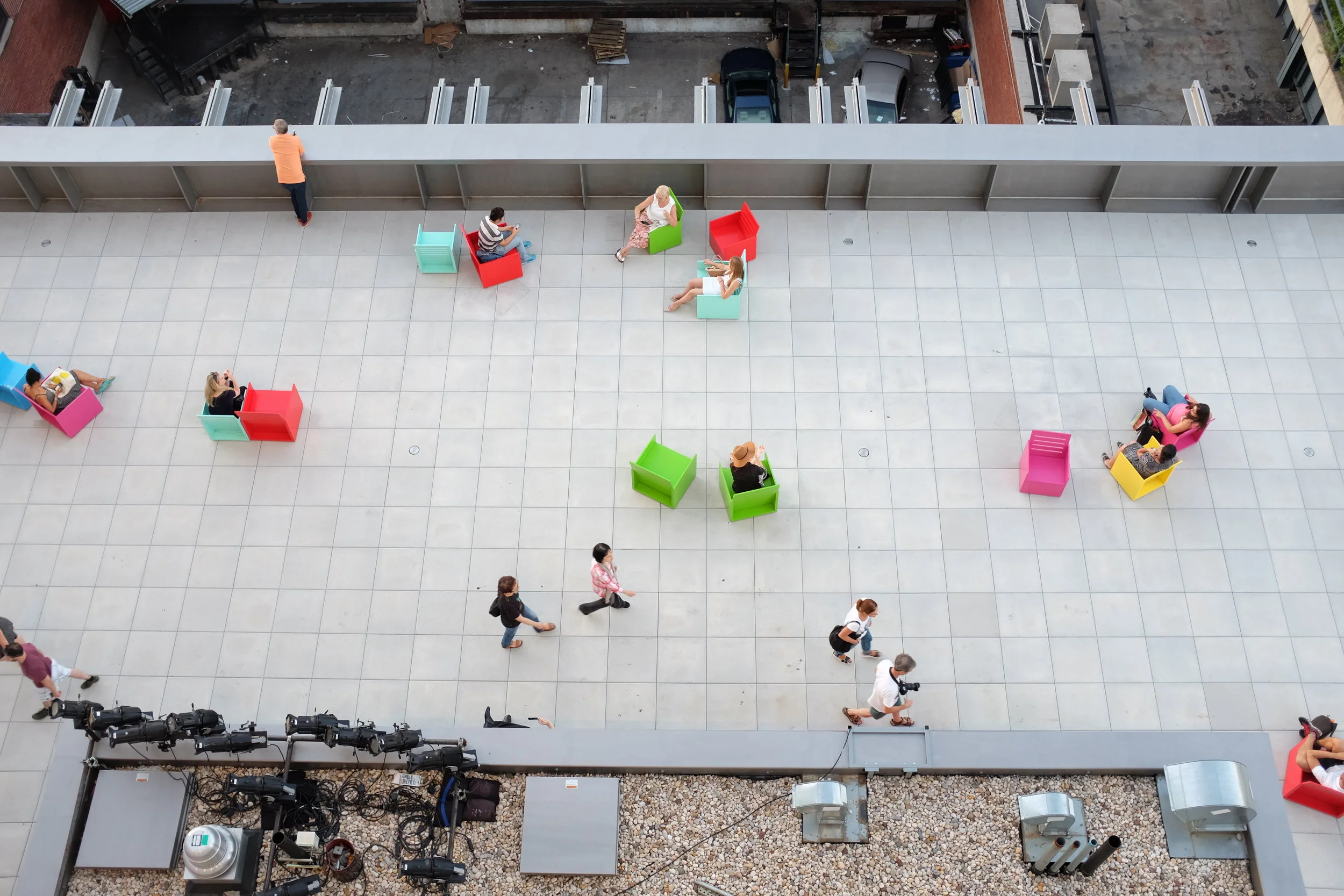 High-angle view of a rooftop terrace at MoMA with colorful chairs and people sitting, walking, and standing. There is a parking lot with cars and a few people in the background, and some professional photographer equipment near the edge.