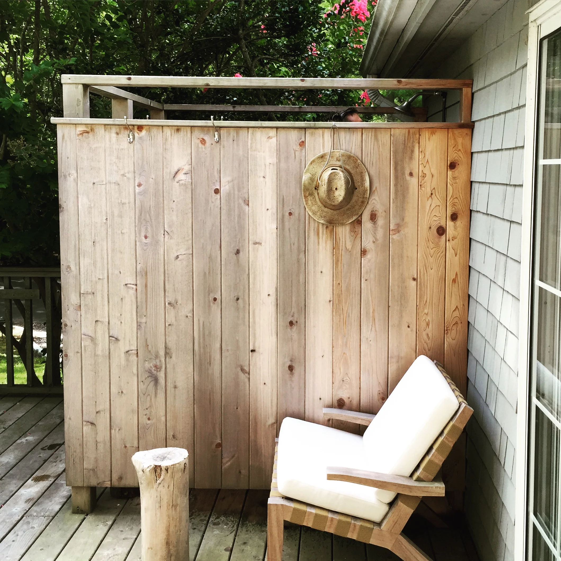 A wooden privacy wall on a deck with a hat hanging on it, a cushioned chair with wooden armrests, and a small tree stump in front. There are trees and greenery visible beyond the wall.