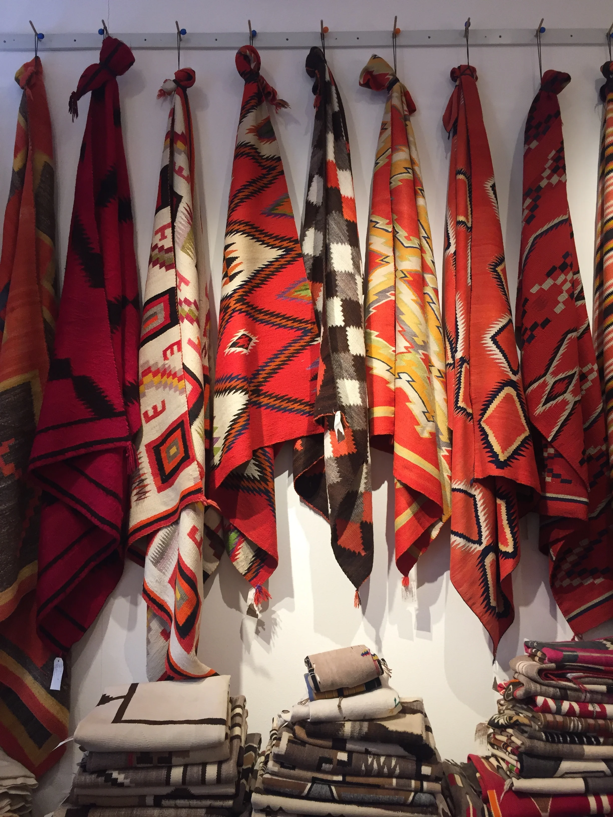 A display of colorful woven blankets or shawls hanging on hooks, featuring vibrant patterns with geometric designs in colors like red, black, white, and beige.