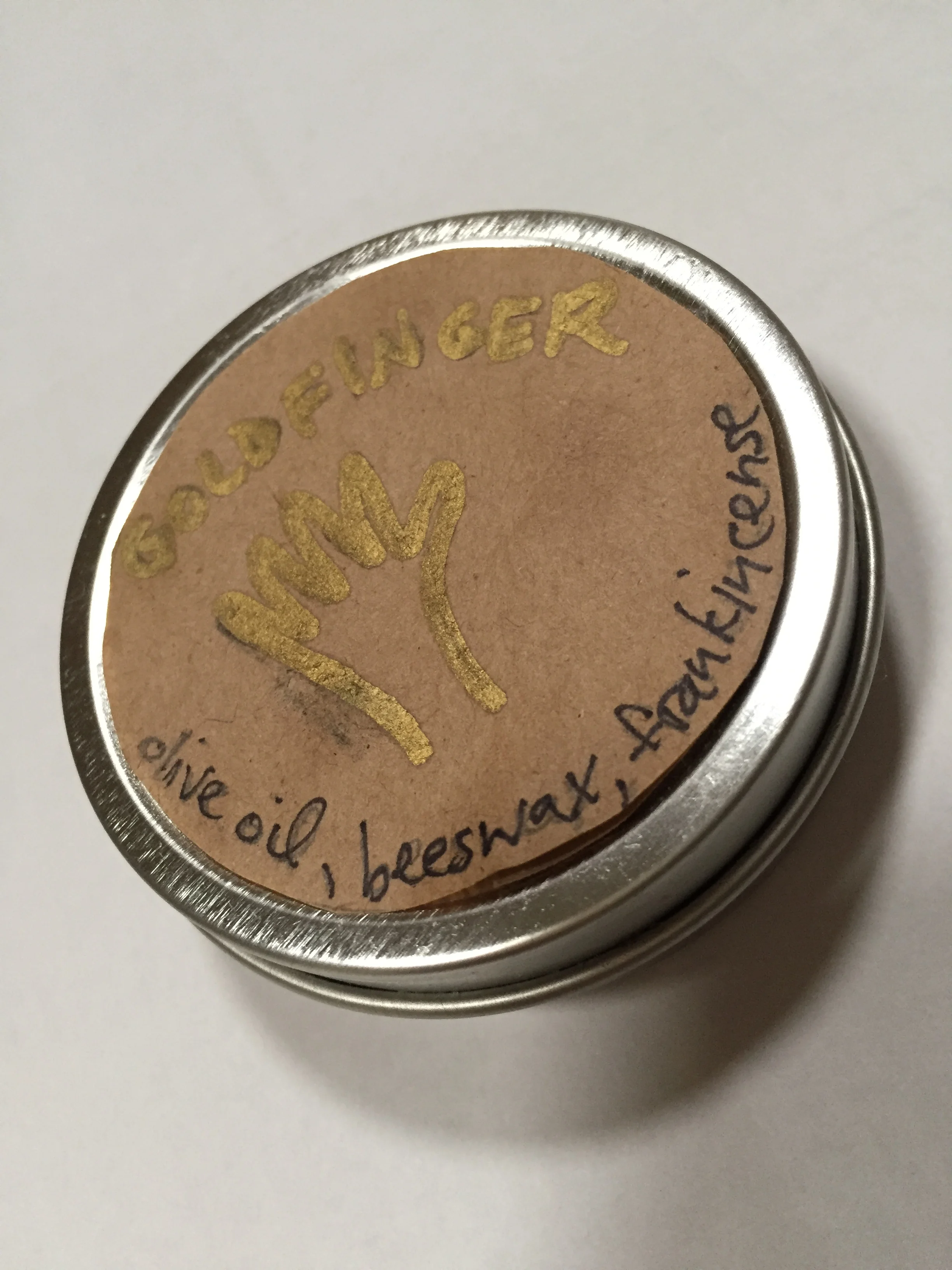 A small round tin container with a brown paper label on the lid. The label has a gold-colored drawing of a hand giving a thumbs-up, with the words 'GOLDFINGER' in gold above it and 'olive oil, beewax, and fairtrade' written in black around the bottom