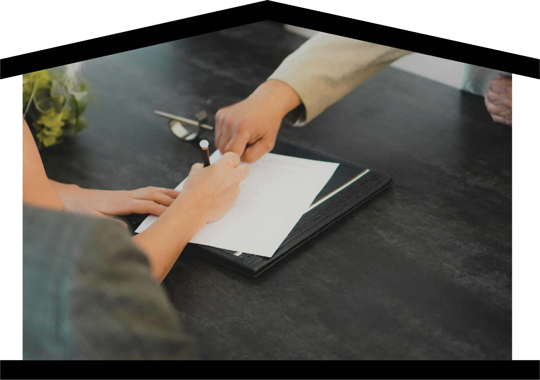 Person signing a document on a desk with a pen, with another person pointing at the paper, and eyeglasses nearby.