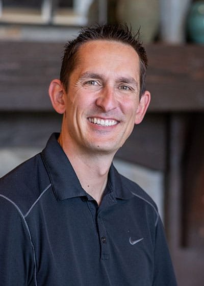 A man with short dark hair smiling, wearing a black Nike polo shirt, standing in front of a blurred background with wooden furniture.  Mark Rainsdon.  OCD, Therapy, Telehealth