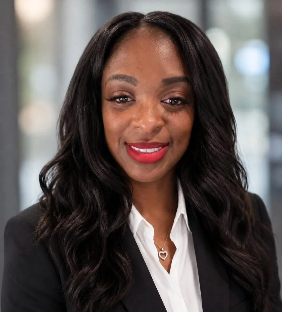 Professional woman with long wavy black hair, wearing a black blazer and white blouse, smiling and showing her teeth, with a heart-shaped pendant necklace.
