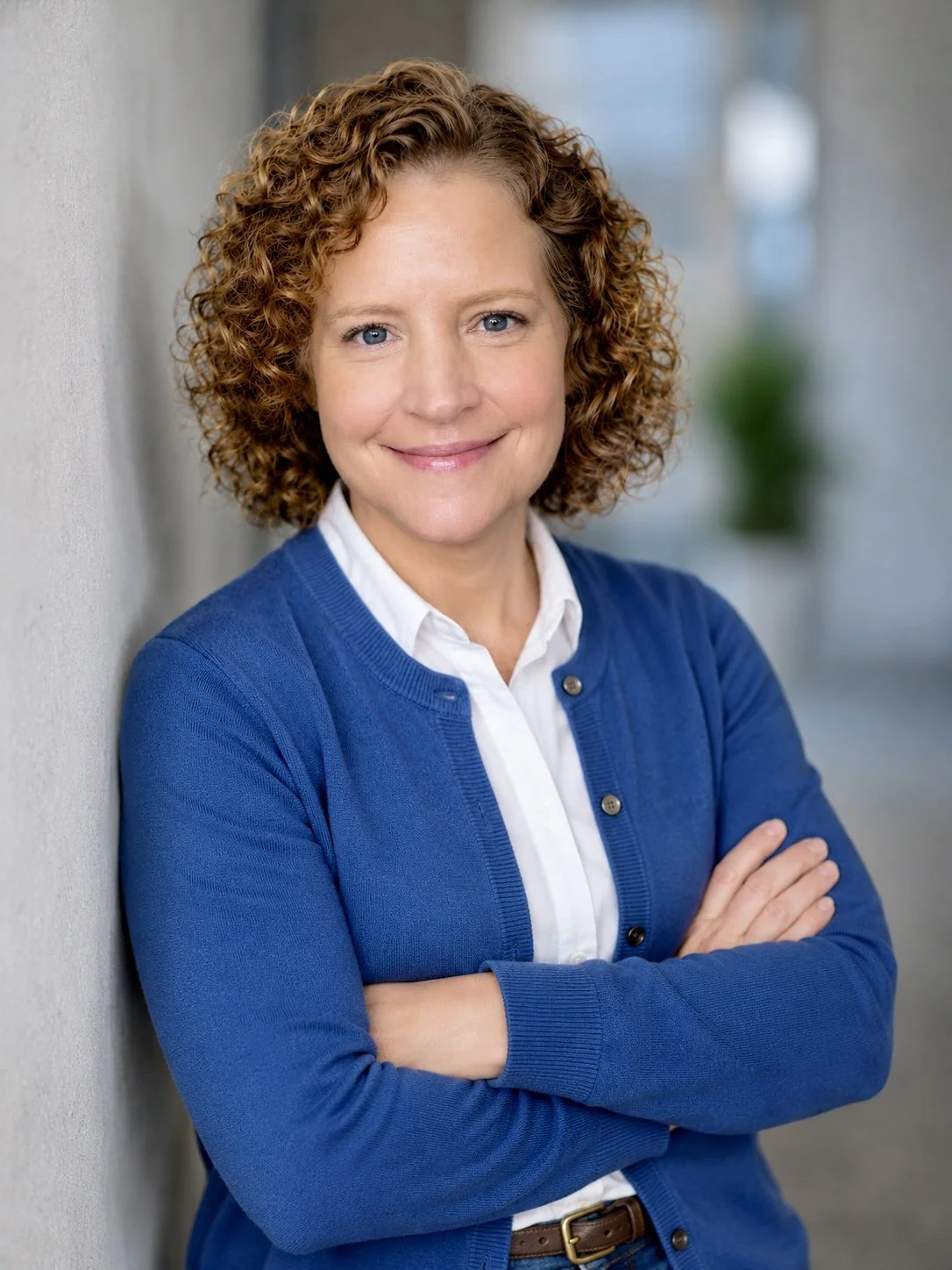 A woman with curly red hair, wearing a white collared shirt and a blue cardigan, smiling while leaning against a wall in an indoor setting.