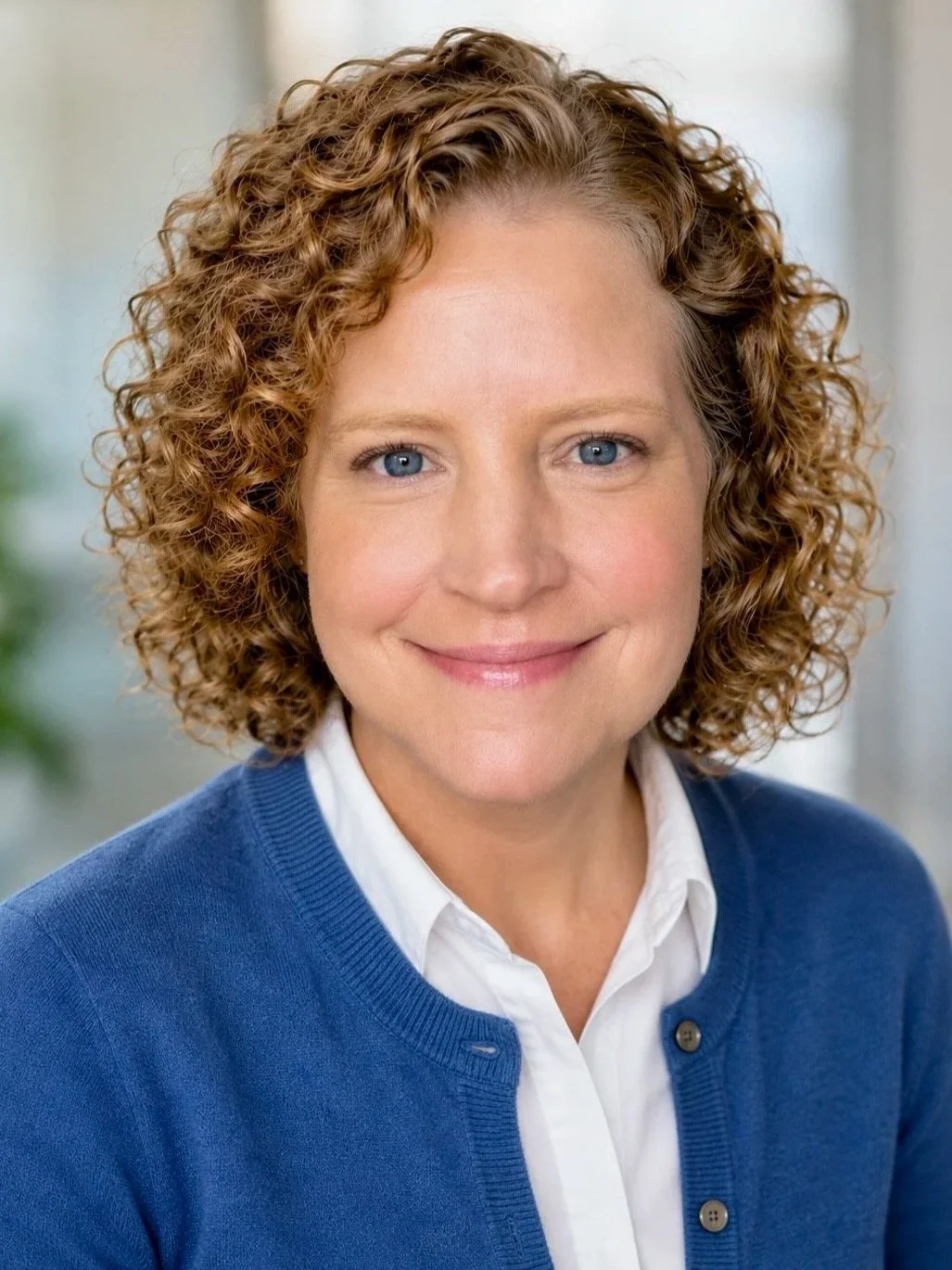 A woman with curly red hair and blue eyes, smiling, wearing a white shirt under a blue cardigan.
