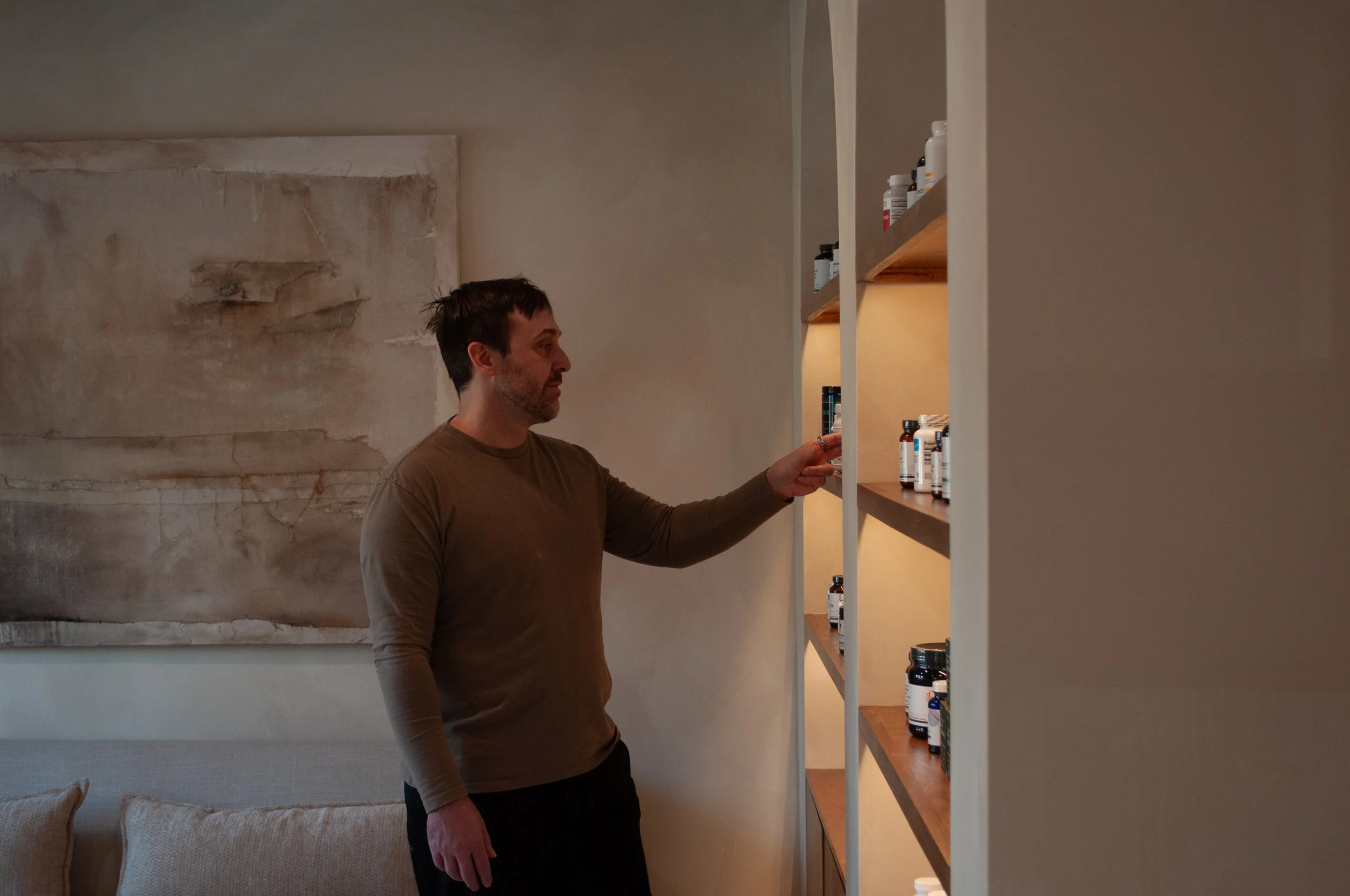 A man stands in a living room reaching for a bottle on a shelf filled with medicine and supplements.
