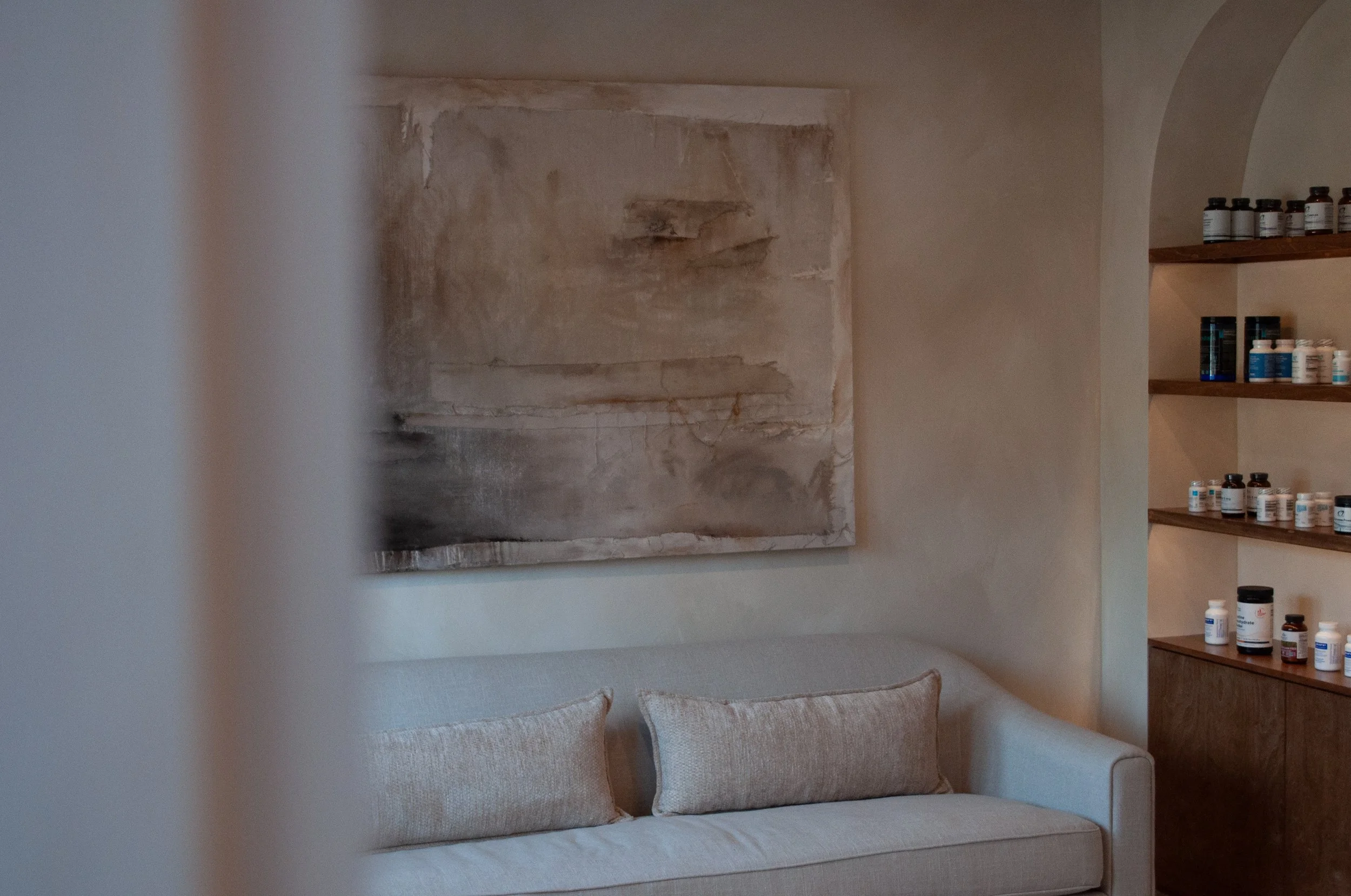 A beige sofa with two cushions in front of an abstract painting on a textured wall, with a wooden shelf filled with bottles on the right.