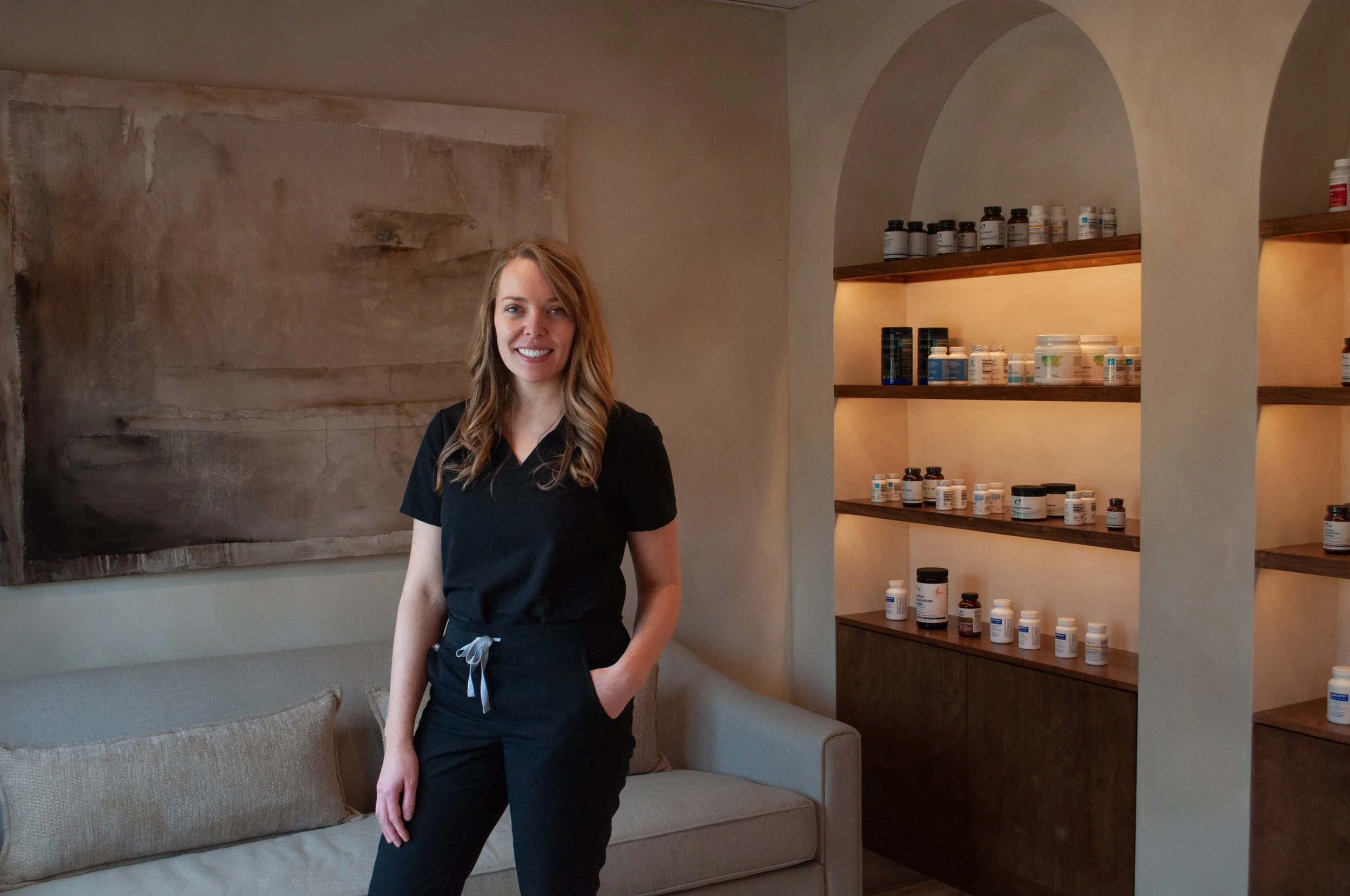 A woman in black scrubs standing in a room with beige walls, a beige couch with cushions, and wooden shelves filled with bottles of supplements or medication.