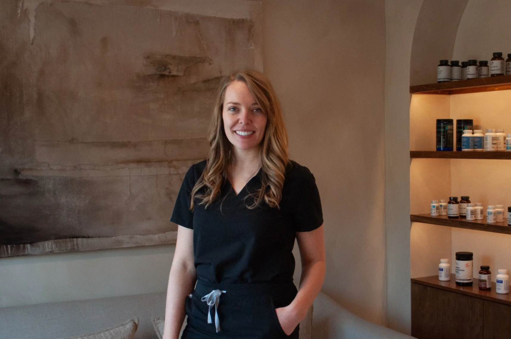 A woman in black scrubs standing in a room with beige walls and a wooden shelf with bottles on it, smiling at the camera.