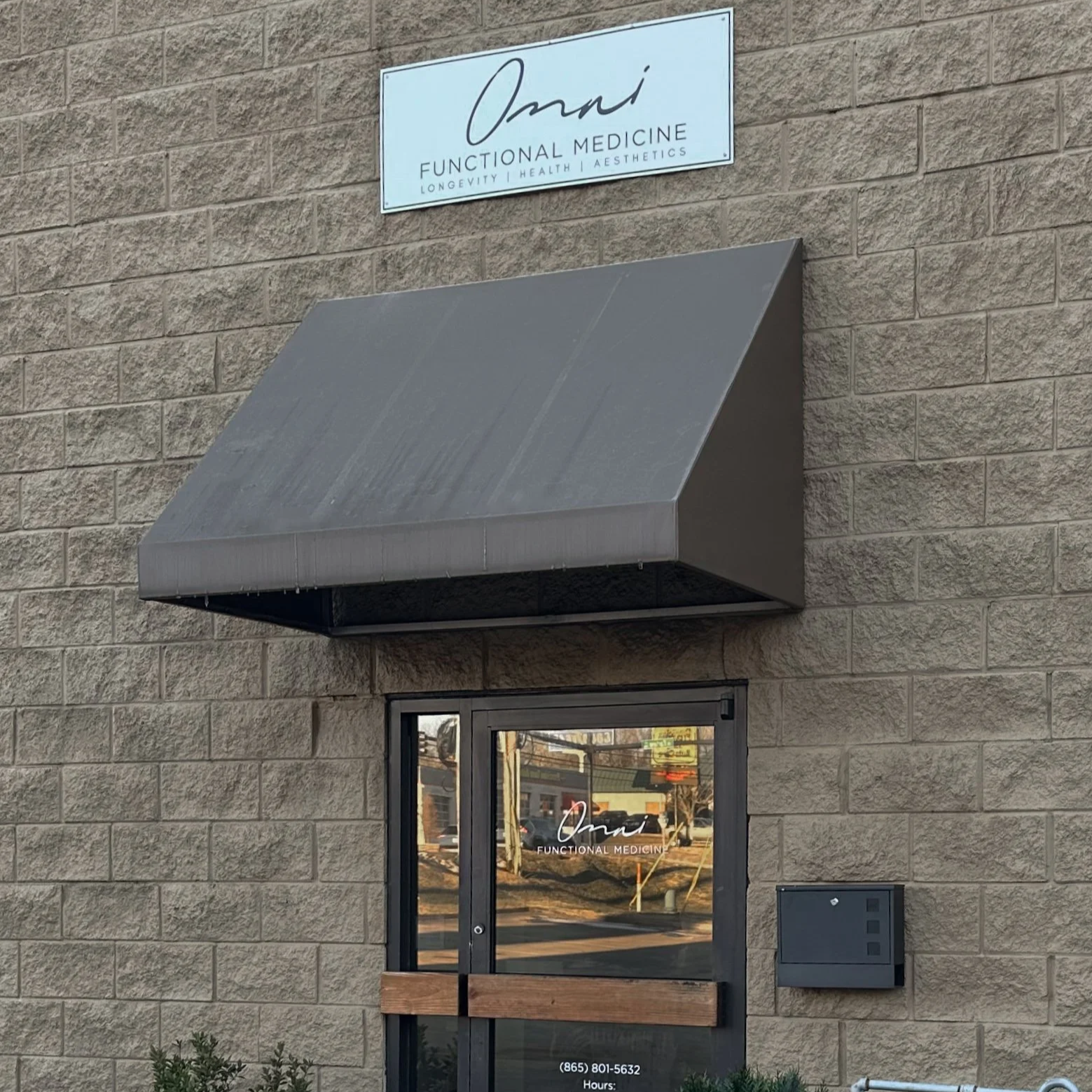 The exterior of a functional medicine clinic called Omn, featuring a sign above the door with the clinic name and services, a metal awning, a glass door with the clinic name and hours, and a mailbox on the wall.