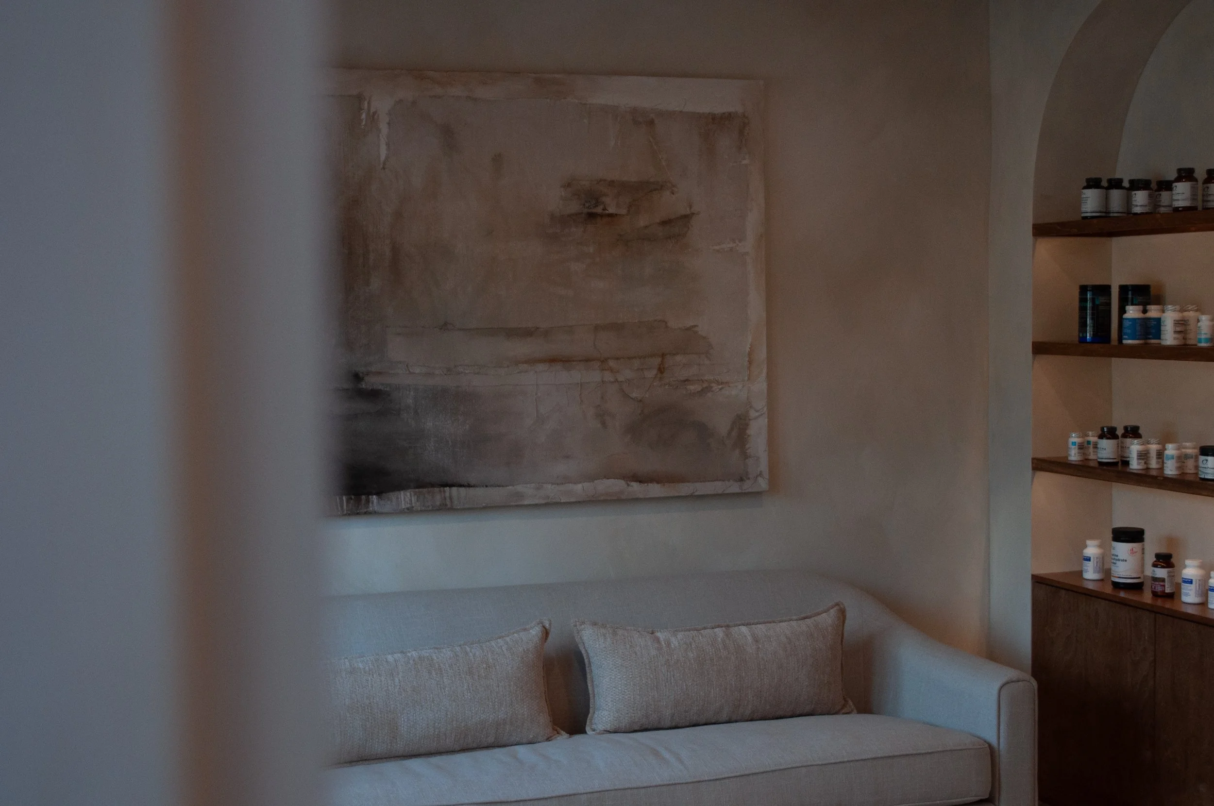 Spa like waiting room with beige sofa, abstract wall art with fabric texture, and wooden shelves with supplement and skincare bottles.