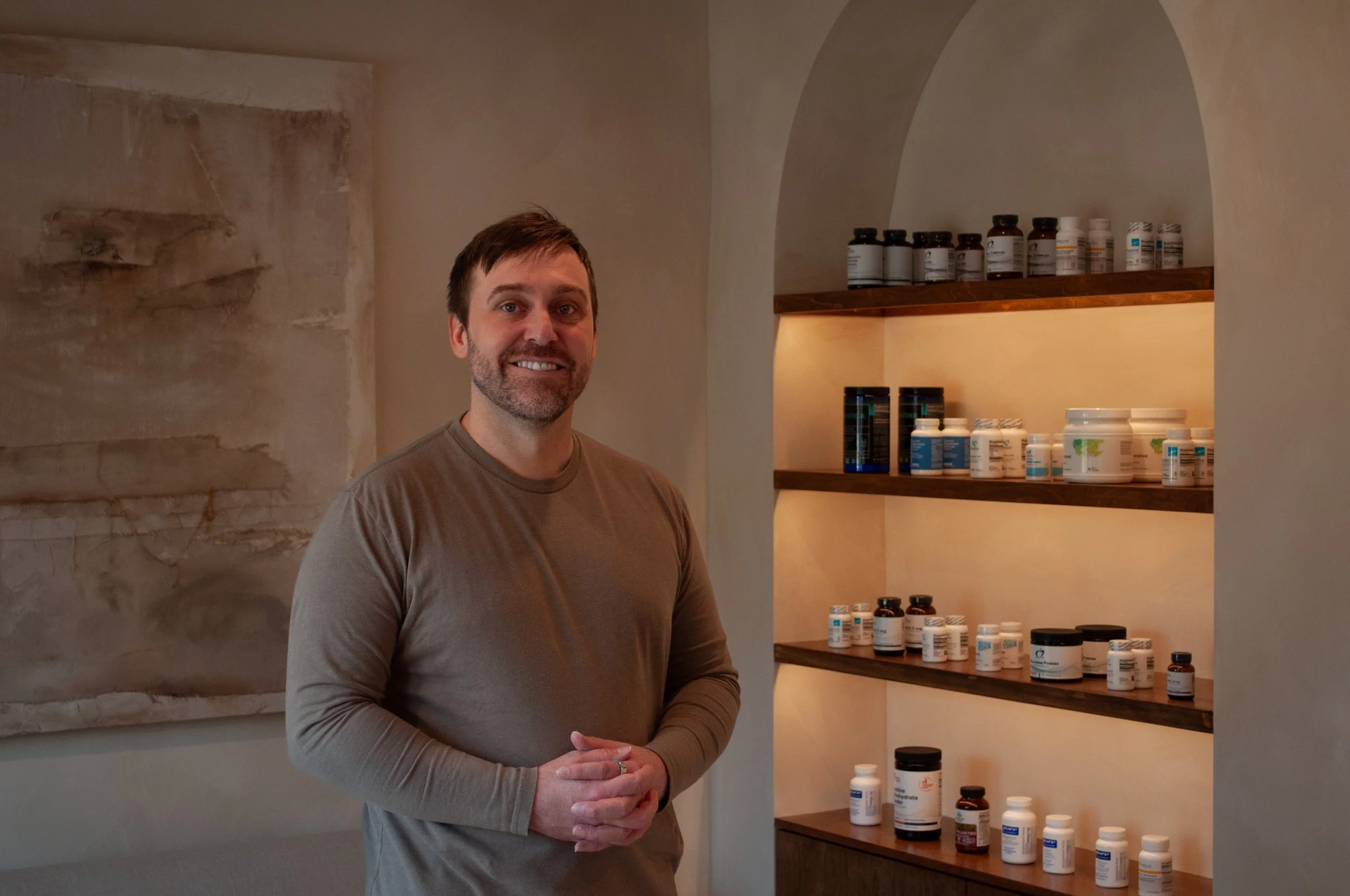A man with a beard and short hair, smiling, stands inside a room with shelves filled with supplement bottles behind him.
